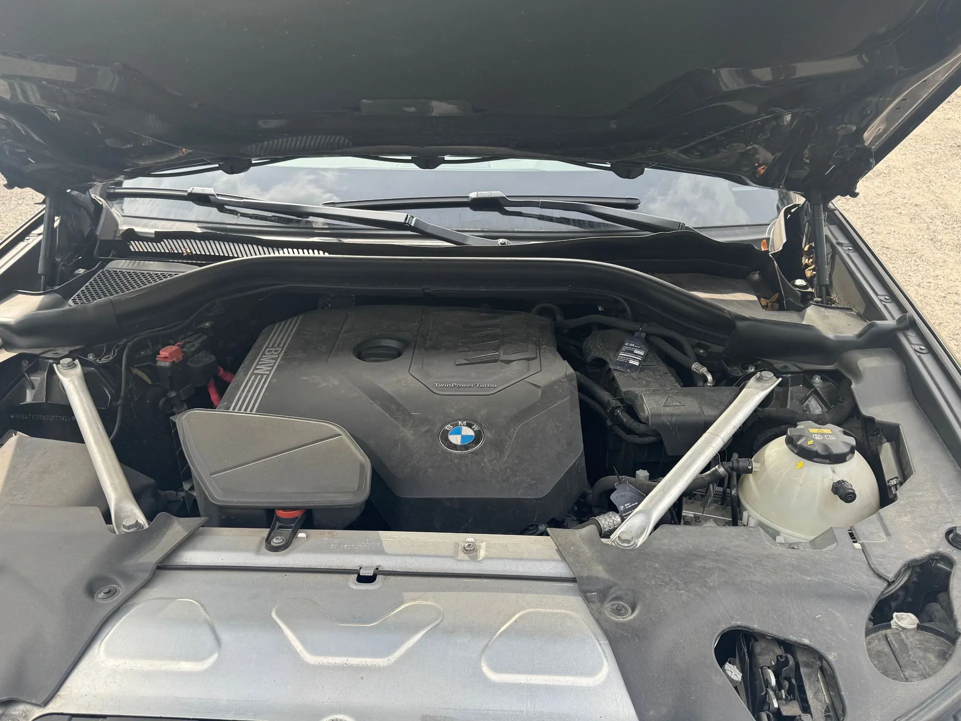 Widok z góry na komorę silnika czarnego samochodu z otwartą maską. Widoczna jest plastikowa osłona silnika z logo BMW oraz napisem 'TwinPower Turbo'. Po bokach znajdują się metalowe rozpórki, a po prawej stronie biały zbiornik z płynem. W tle widać przednią szybę i wycieraczki.