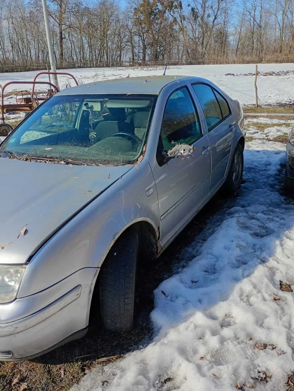 Srebrny samochód osobowy typu sedan zaparkowany na śnieżnej powierzchni. Pojazd ma uszkodzony boczny lusterko i ślady zużycia. W tle widoczne są bezlistne drzewa, fragment czerwonego sprzętu rolniczego oraz pole pokryte śniegiem. Niebo jest jasne i bezchmurne.
