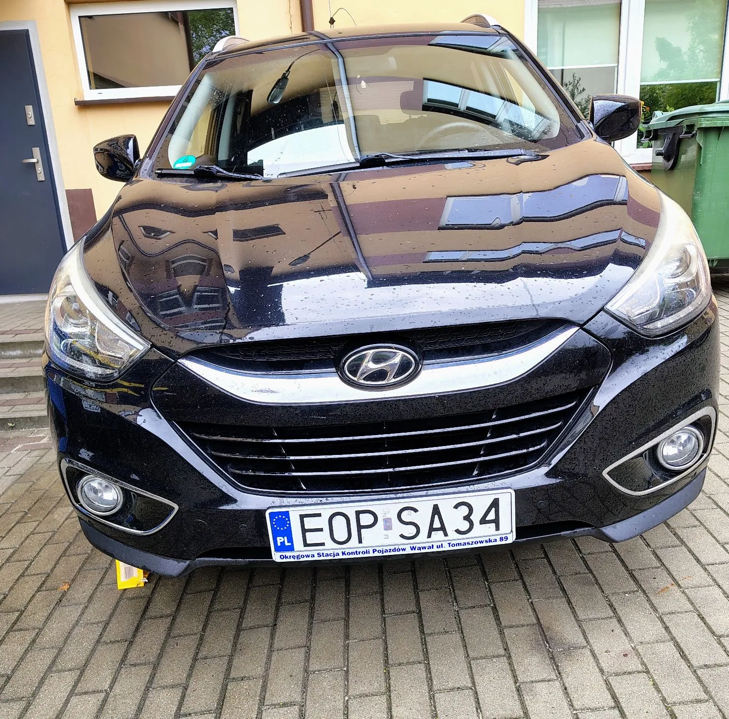 Czarny samochód marki Hyundai zaparkowany przodem do obiektywu na kostce brukowej. Na masce widoczne są krople wody. Samochód posiada polską tablicę rejestracyjną o numerze EOP SA34. Poniżej numeru rejestracyjnego znajduje się napis: 'Okręgowa Stacja Kontroli Pojazdów Wawał ul. Tomaszowska 89'. W tle widoczny jest fragment żółtego budynku z oknem oraz zielony kontener na odpady.