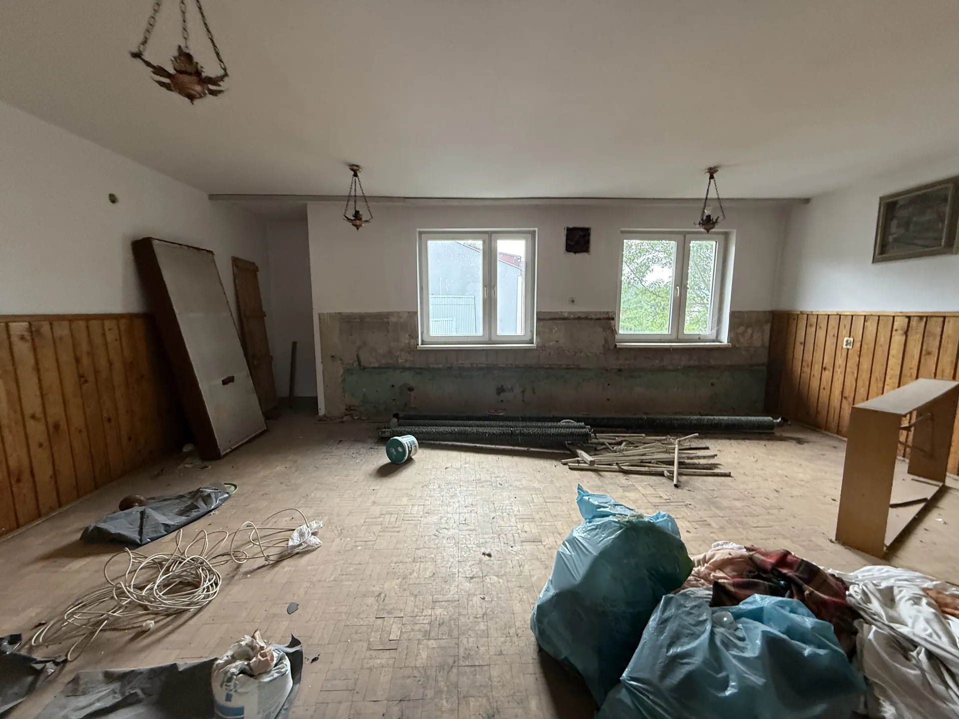 Pomieszczenie w trakcie remontu z podłogą z kafelkami, na której leżą nieporządki: niebieskie worki na śmieci, zwinięte przewody, deski, wiadro i materiały budowlane. Ściany częściowo obite deskami, z widocznymi uszkodzeniami i odsłoniętymi fragmentami muru pod oknami. W tle dwa okna z widokiem na zewnątrz, trzy wiszące lampy sufitowe, drzwi po lewej stronie oraz rama obrazu na prawej ścianie. Sufit biały, podłoga brudna i zniszczona.