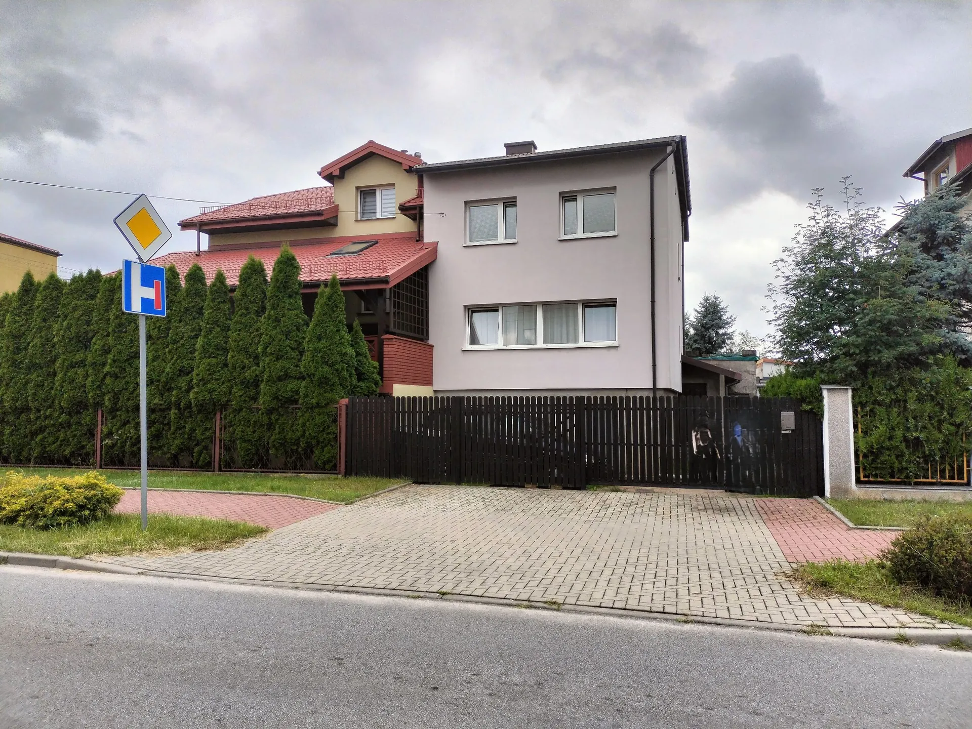 Dwupiętrowy dom jednorodzinny z jasnoszarą elewacją i czerwonym dachem. Przed domem znajduje się ciemnobrązowy, drewniany płot z bramą oraz podjazd wyłożony szarą kostką brukową. Po lewej stronie posesji rośnie rząd wysokich, gęstych tuj, a obok nich stoi znak drogowy z niebieskim symbolem parkingu i żółtym rombem powyżej. Niebo jest zachmurzone.