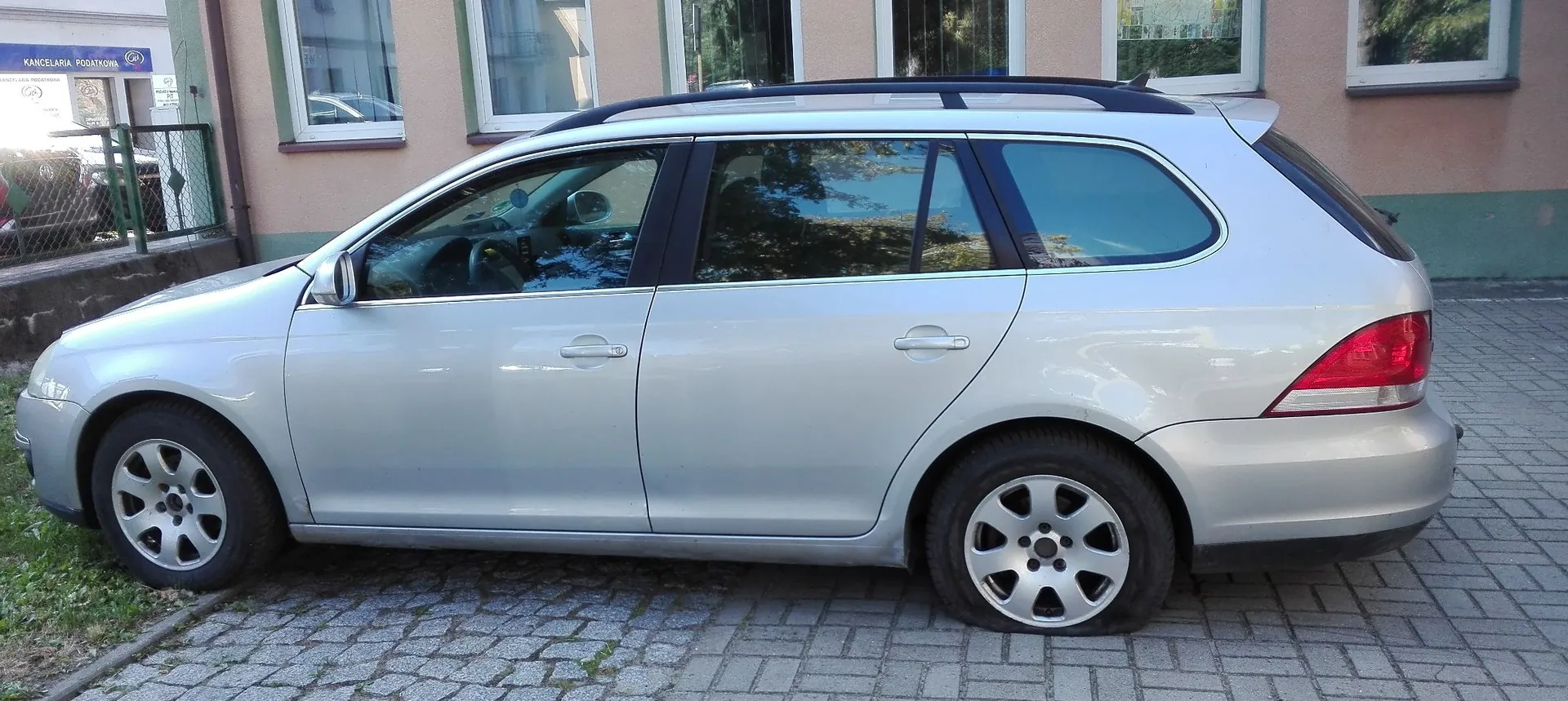 Srebrny samochód osobowy typu kombi zaparkowany bokiem na kostce brukowej przed budynkiem. W tle widoczna ściana budynku z oknami oraz fragment zielonego ogrodzenia z tabliczką z napisem 'KANCELARIA PODATKOWA'.