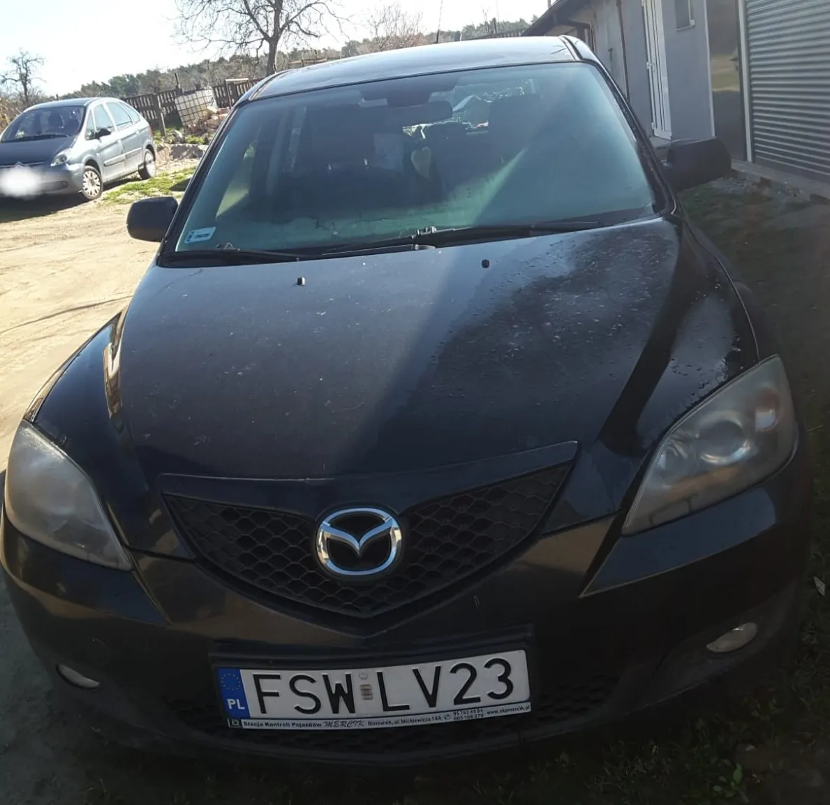Czarny samochód marki Mazda z przodu, z widoczną tablicą rejestracyjną FSW LV23 z kodem PL po lewej stronie. Na tle srebrny samochód zaparkowany na podwórku, budynek z bramą garażową oraz drzewa. Słońce oświetla scenę, na masce widoczne są krople wody.