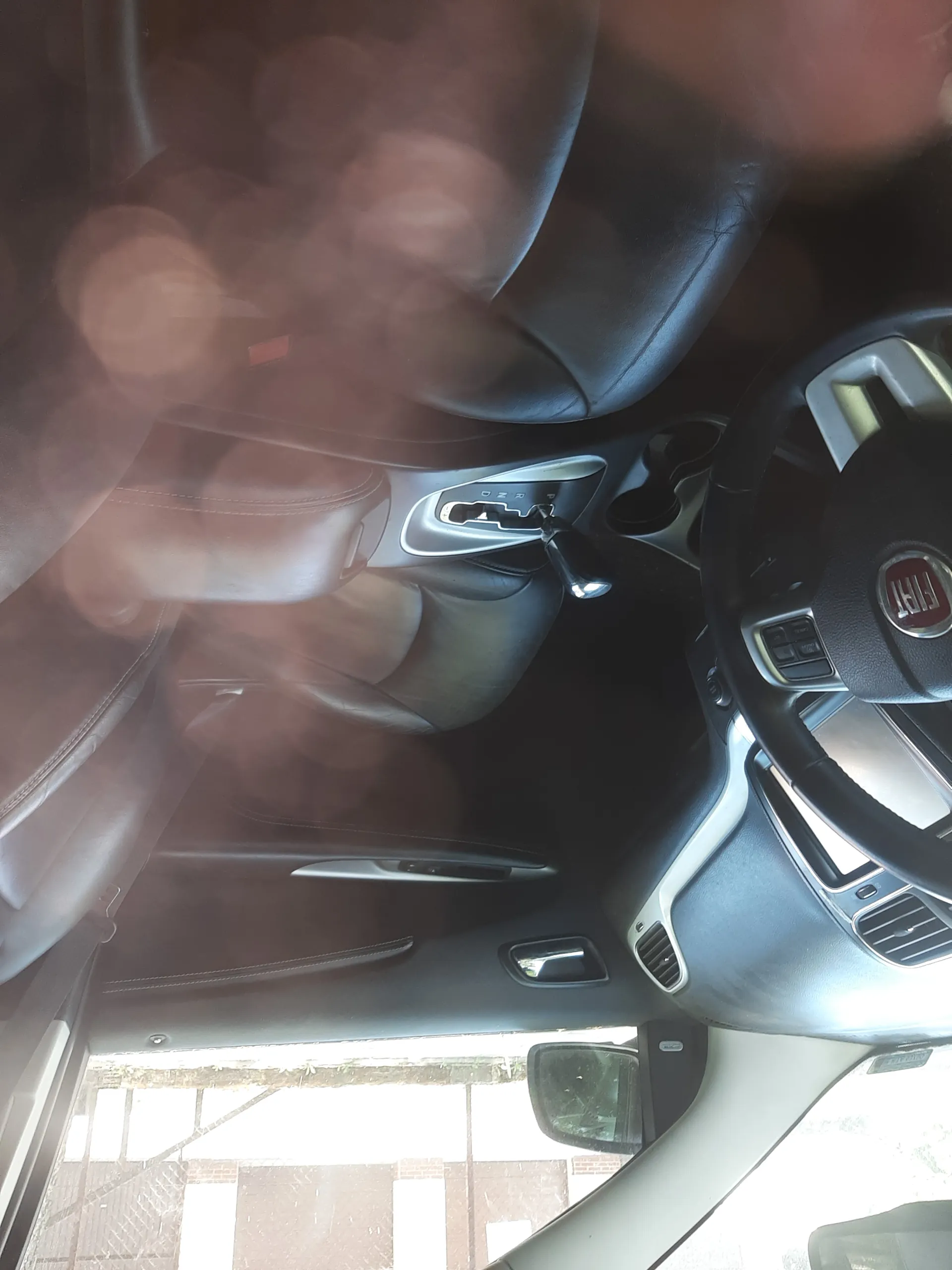 Wnętrze samochodu marki Fiat. Widoczna kierownica z logo FIAT, deska rozdzielcza z wentylacjami, skrzynia biegów z oznaczeniami P R N D, czarne i brązowe fotele, fragment drzwi z lusterkiem bocznym oraz widok na zewnątrz przez okno. Na szybie widoczne są plamy lub rozmazane refleksy świetlne.