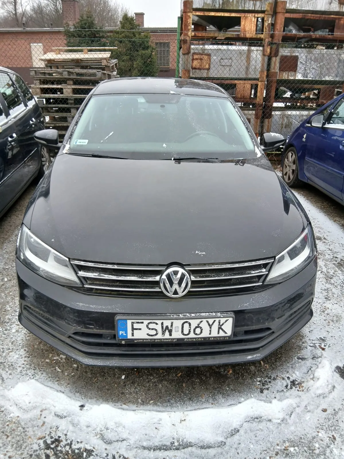 Czarny samochód marki Volkswagen z tablicą rejestracyjną FSW 06YK (PL) na przodzie, zaparkowany na obszarze z resztkami śniegu. Po lewej stronie czarny samochód, po prawej niebieski. W tle siatka ogrodzeniowa, stosy palet, ceglany budynek z kominami oraz drzewa.