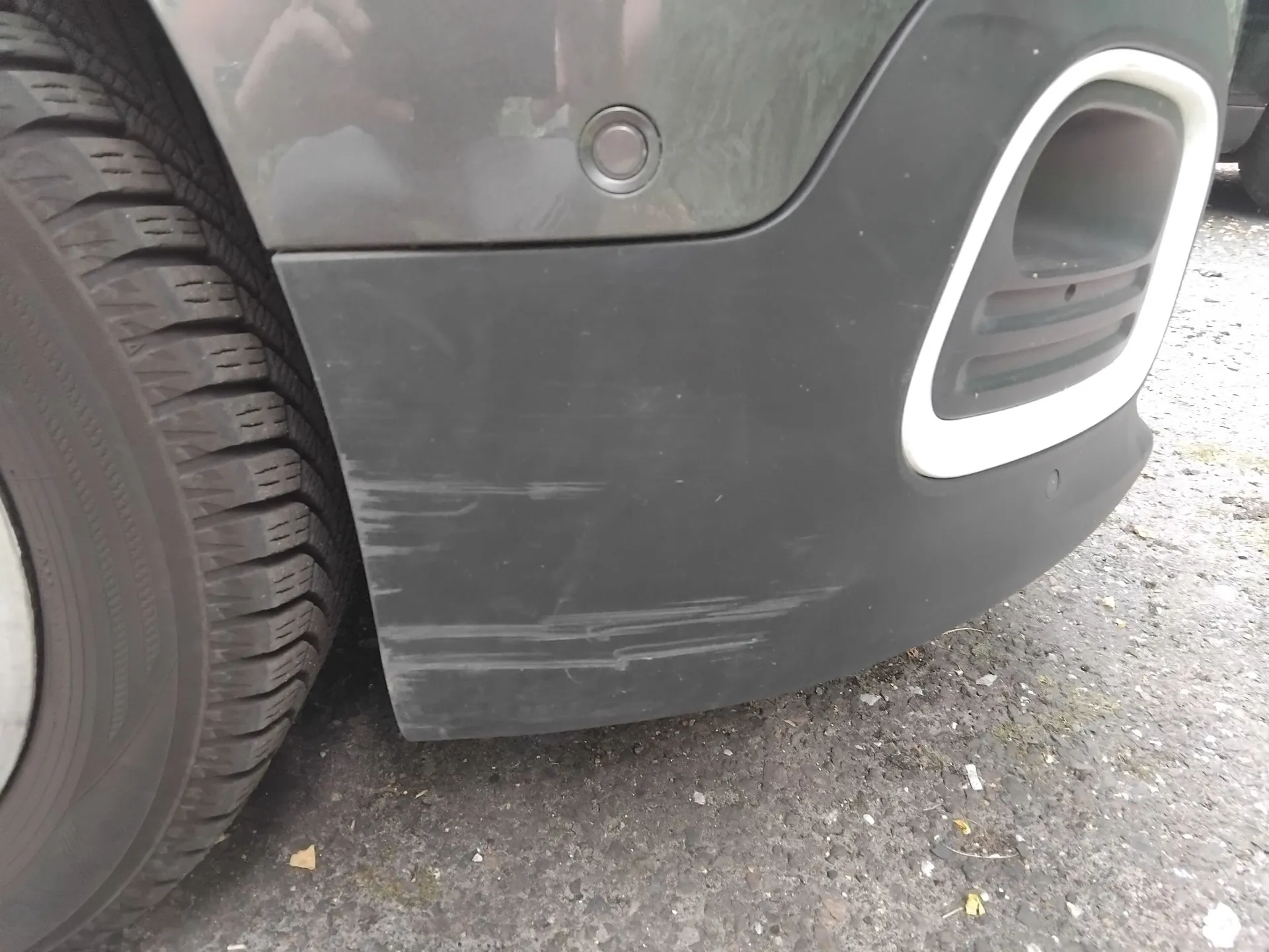 Zbliżenie na tylny zderzak ciemnoszarego samochodu. Na czarnym, plastikowym elemencie zderzaka widoczne są liczne, poziome zadrapania i otarcia. Po prawej stronie znajduje się biała obramowana wnęka wydechu. Po lewej stronie widoczny jest fragment opony i felgi samochodu. Pojazd stoi na asfaltowej nawierzchni.