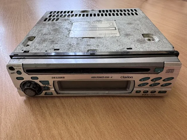 Srebrne radio samochodowe marki Clarion, model DB328RB, leżące na drewnianym blacie. Urządzenie jest zakurzone. Na przednim panelu widnieją napisy: 'DB328RB', 'HIGH POWER 45W x 4', 'Clarion' oraz 'R-D-S / EON'. Po lewej stronie znajduje się pokrętło głośności, a po prawej zestaw przycisków sterujących. Na górnej obudowie widoczna jest biała naklejka z danymi technicznymi i numerem seryjnym.