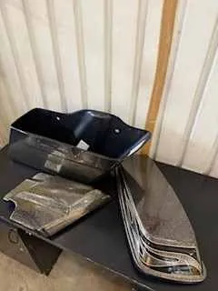 Na czarnym stole znajdują się trzy elementy samochodowe: czarna plastikowa obudowa, metalowa część z przezroczystą osłoną oraz druga metalowa część z kilkoma poziomymi liniami. Tło stanowią pionowe deski drewniane. Brak czytelnego tekstu.