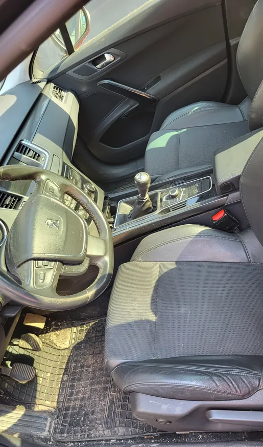 Wnętrze samochodu z perspektywy kierowcy. Widoczna jest czarna skórzana tapicerka foteli, kierownica z logo marki Peugeot, lewarek zmiany biegów w konsoli środkowej oraz czarna gumowa mata na podłodze. W tle widoczny jest panel drzwi kierowcy oraz fragment deski rozdzielczej.