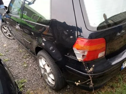 Czarny samochód widziany od tyłu, zaparkowany na żwirowej powierzchni z fragmentem trawy. Widoczne są czerwone i pomarańczowe światła tylnego, srebrna felga oraz fragment tablicy rejestracyjnej. Po lewej stronie częściowo widoczny inny pojazd. W tle odbicie zielonego obiektu w szybie oraz sucha gałązka przy zderzaku.