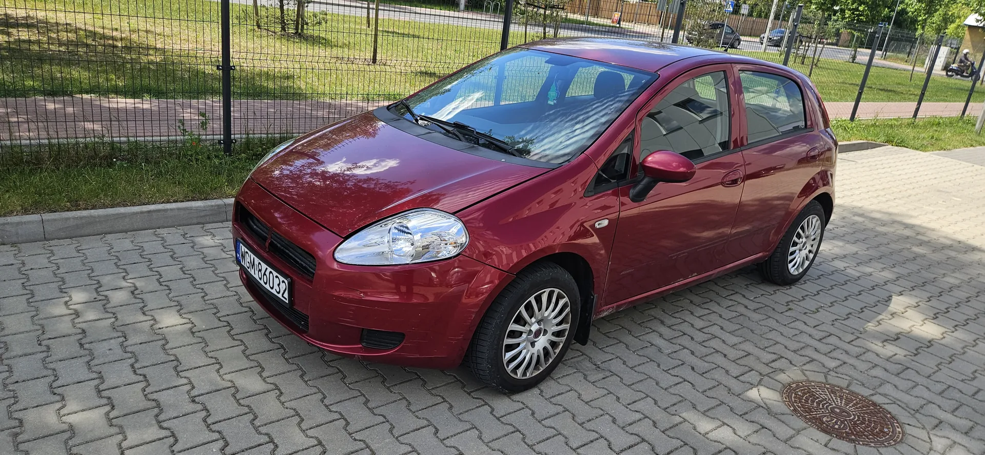 Czerwony samochód osobowy typu hatchback zaparkowany na brukowanej powierzchni. Widoczna tablica rejestracyjna WGM 86032. W tle metalowe ogrodzenie, trawnik, chodnik oraz drzewa. Na prawo od samochodu znajduje się okrągła klapa studzienki. W oddali widoczne są inne pojazdy i droga.
