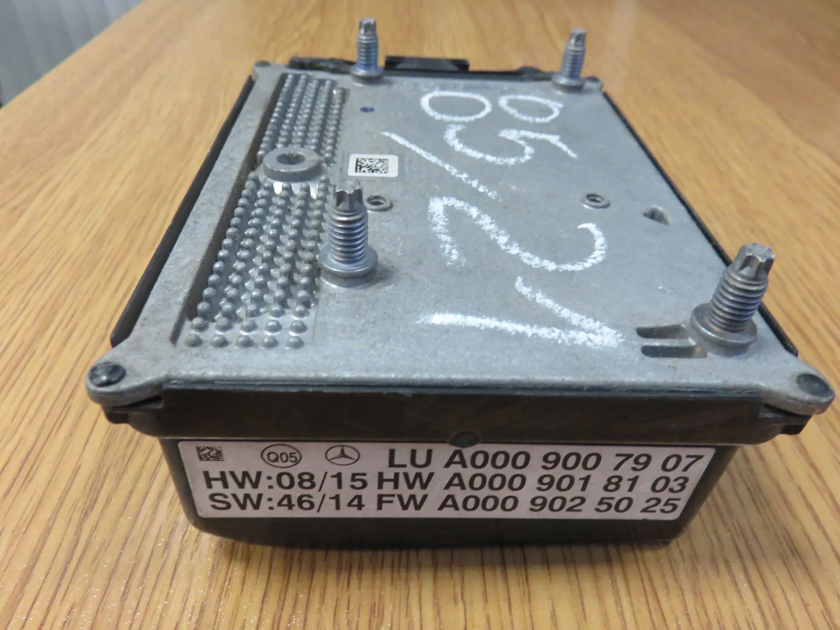 Metalowa obudowa modułu elektronicznego leżąca na drewnianym blacie. Na górnej części obudowy znajdują się cztery wystające śruby mocujące oraz białe, odręczne napisy. Na bocznej ściance naklejona jest etykieta z tekstem: '(Q05) LU A000 900 79 07 HW:08/15 HW A000 901 81 03 SW:46/14 FW A000 902 50 25' oraz logo marki Mercedes-Benz.