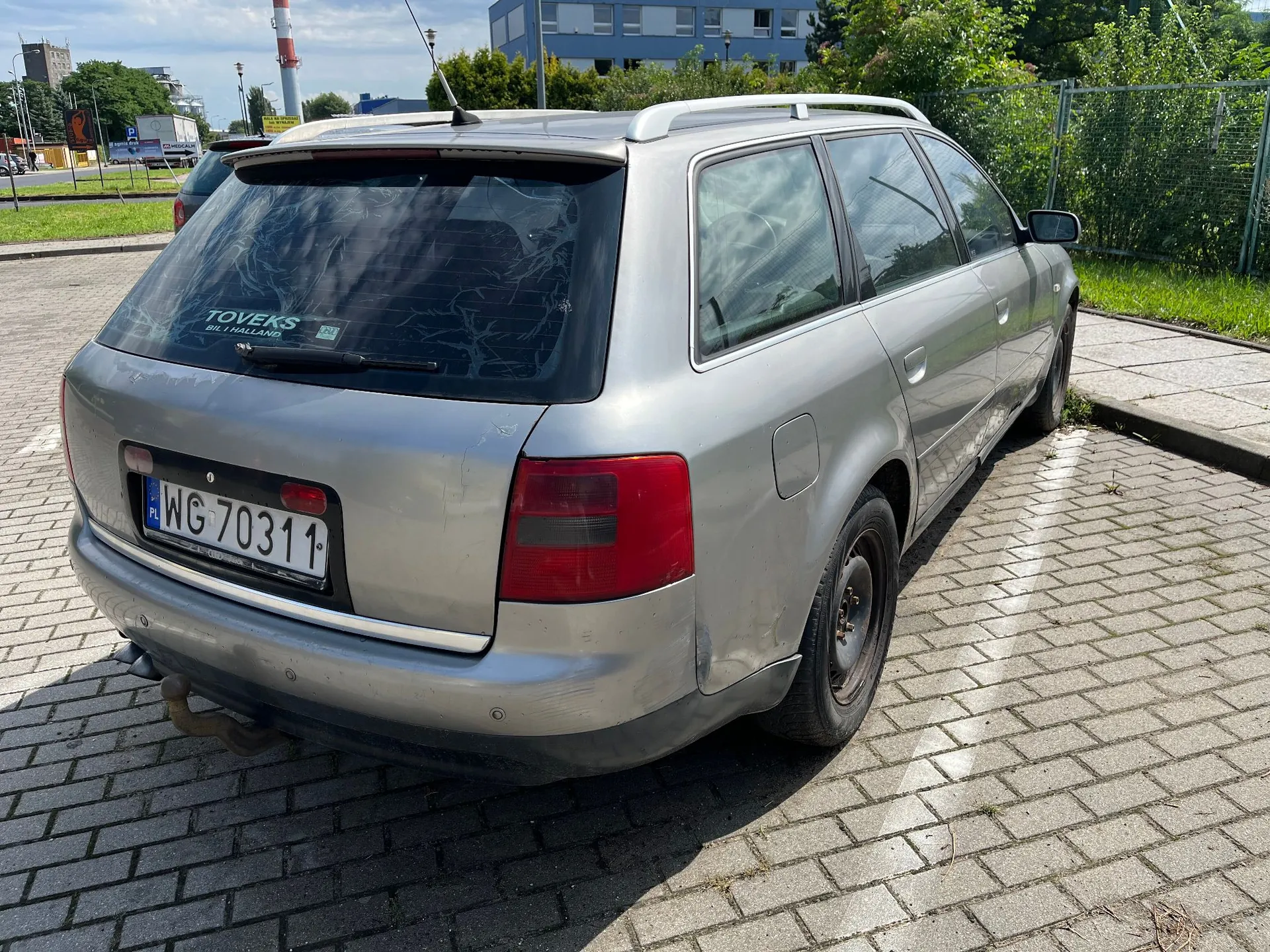 Srebrny samochód typu kombi zaparkowany tyłem na kostce brukowej. Na tylnej szybie znajduje się naklejka z napisem 'TOVEKS BIL I HOLLAND'. Tablica rejestracyjna samochodu to 'WG 70311'. W tle widoczne są budynki, zieleń oraz ogrodzenie.