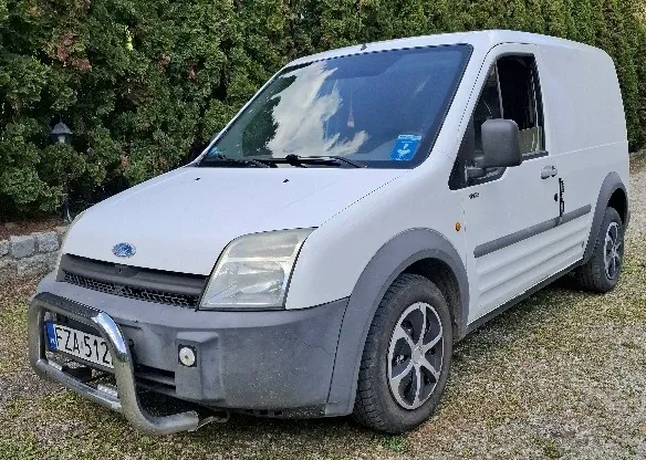 Biały samochód dostawczy marki Ford zaparkowany na trawie z drobnym żwirem. Widoczna tablica rejestracyjna FZA 512, przedni zderzak z metalowym uchwytem, reflektory i logo Ford na masce. Tło stanowią zielone krzewy i kamienny mur. Na przedniej szybie odbicie nieba z chmurami. Samochód ma szare elementy na zderzaku i nadkółach.
