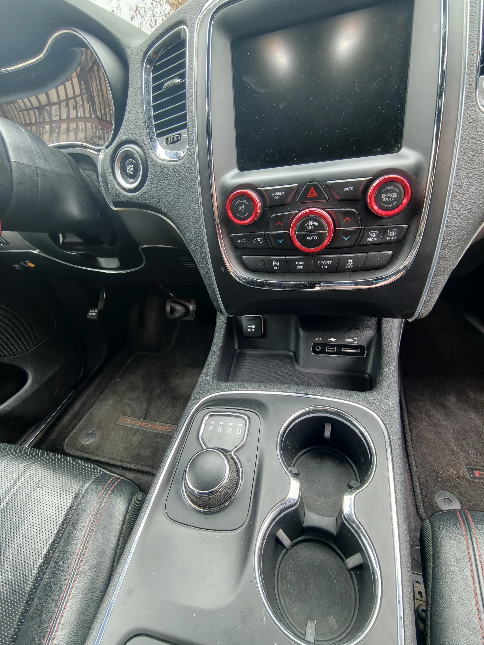 Wnętrze samochodu marki Dodge z widokiem na deskę rozdzielczą i centralną konsolę. Widoczne są czarne siedzenia z czerwonym przeszyciem, kierownica po lewej stronie, ekran multimedialny, przyciski sterujące z czerwonymi akcentami (w tym napisy: AUTO, SPORT, OFF, BACK, BROWSE, ENTER, A/C, FRONT, REAR, P, eco), pokrętło zmiany biegów z oznaczeniem PRND, uchwyty na napoje oraz dywaniki z napisem DODGE. W tle przez przednią szybę widoczne są budynki.