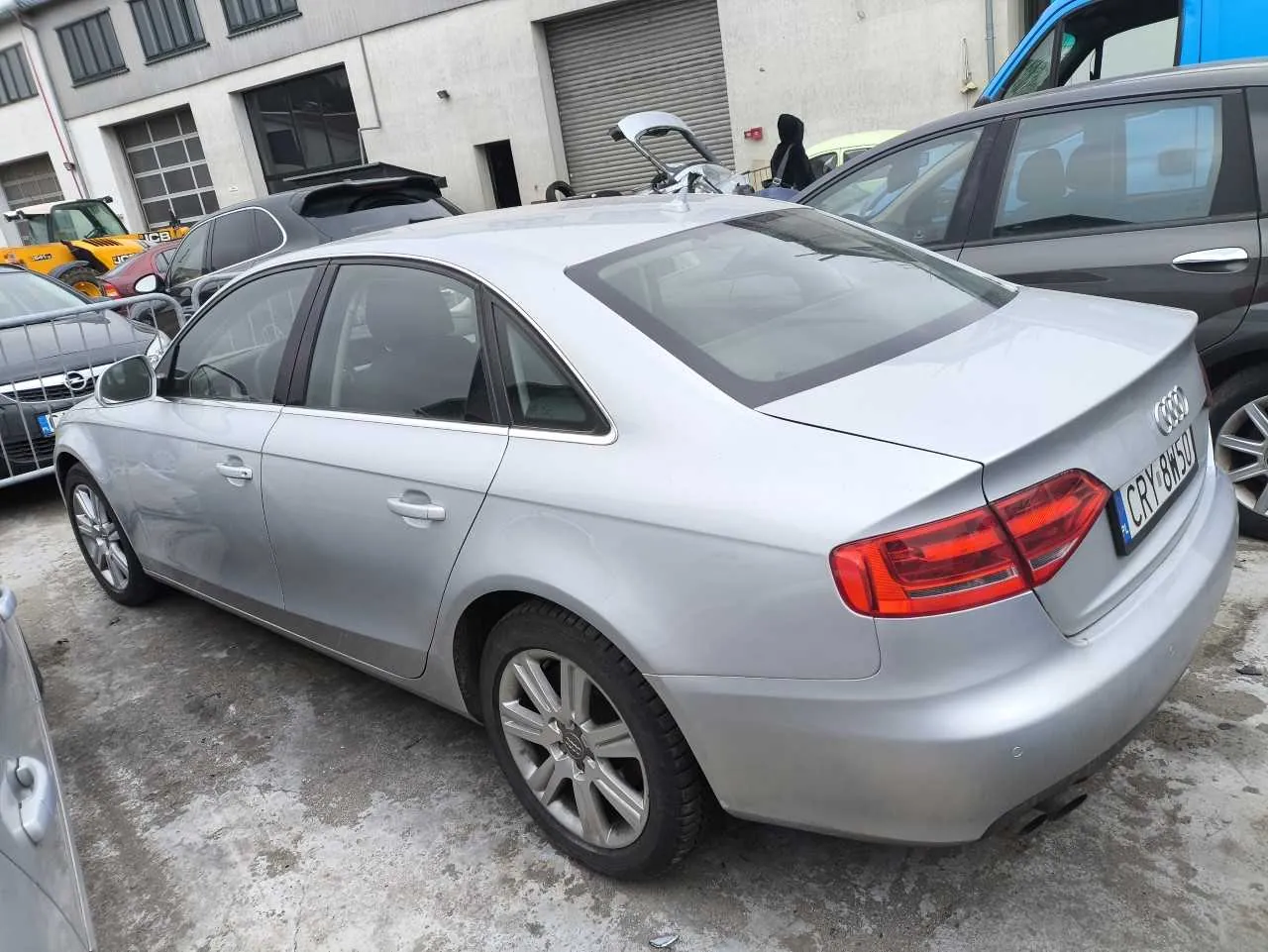 Srebrny samochód Audi sedan z widoczną tablicą rejestracyjną CRV 8W50, zaparkowany na betonowej powierzchni. W tle budynki z zamkniętymi bramami garażowymi, inne pojazdy (m.in. ciemnoszary SUV, żółty pojazd budowlany JCB) oraz osoba w czarnej kurtce. Samochód posiada czarny dach panoramiczny i srebrne felgi.