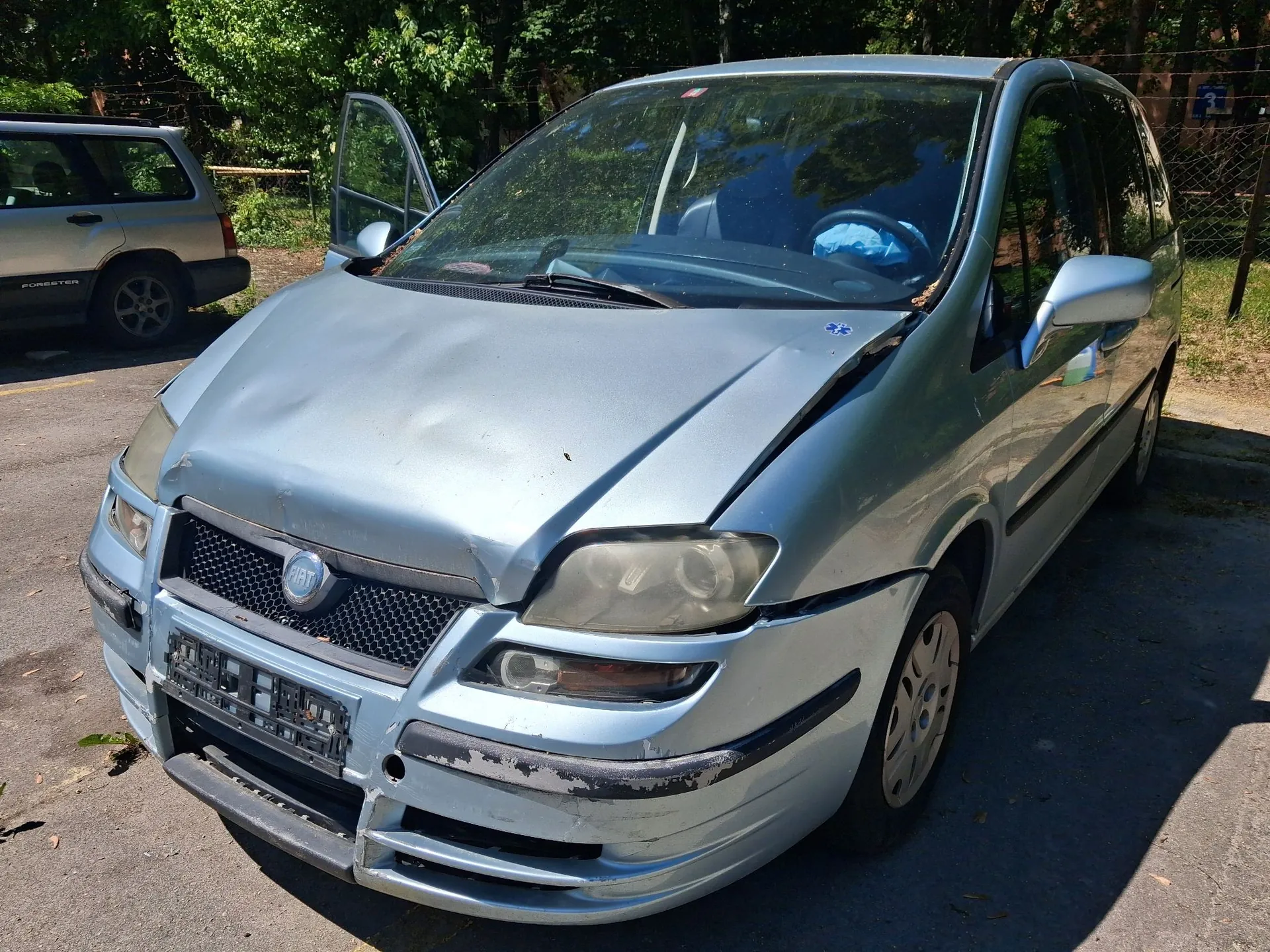 Srebrny samochód typu minivan z widocznymi uszkodzeniami karoserii z przodu, w tym wgnieciona maska i pęknięty zderzak. Pojazd posiada logo FIAT na grillu. Drzwi kierowcy są otwarte. Samochód zaparkowany jest na asfaltowej nawierzchni na tle zielonych drzew.