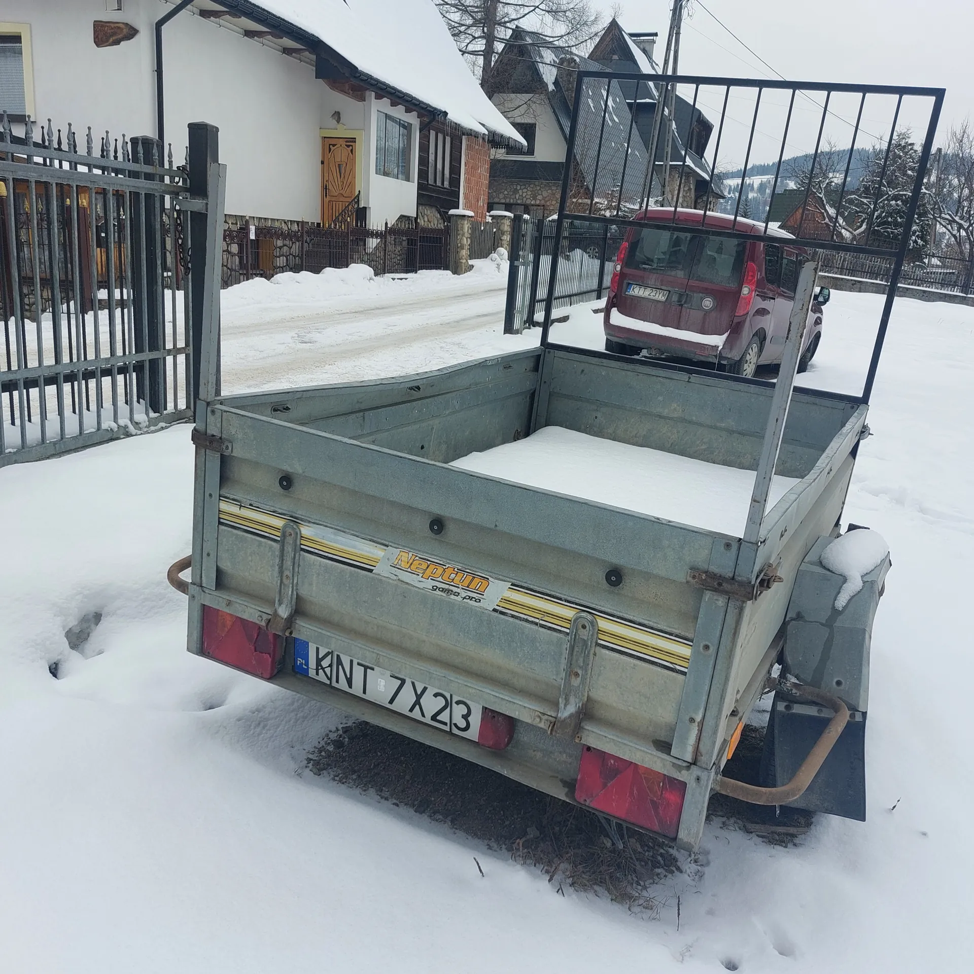 Szary przyczepa z żółtymi paskami, tablicą rejestracyjną KNT 7X23 i napisem 'Neptun'. Za przyczepą czerwony samochód. W tle domy z dachami pokrytymi śniegiem, metalowa brama i śnieżna sceneria. Całość pokryta śniegiem.