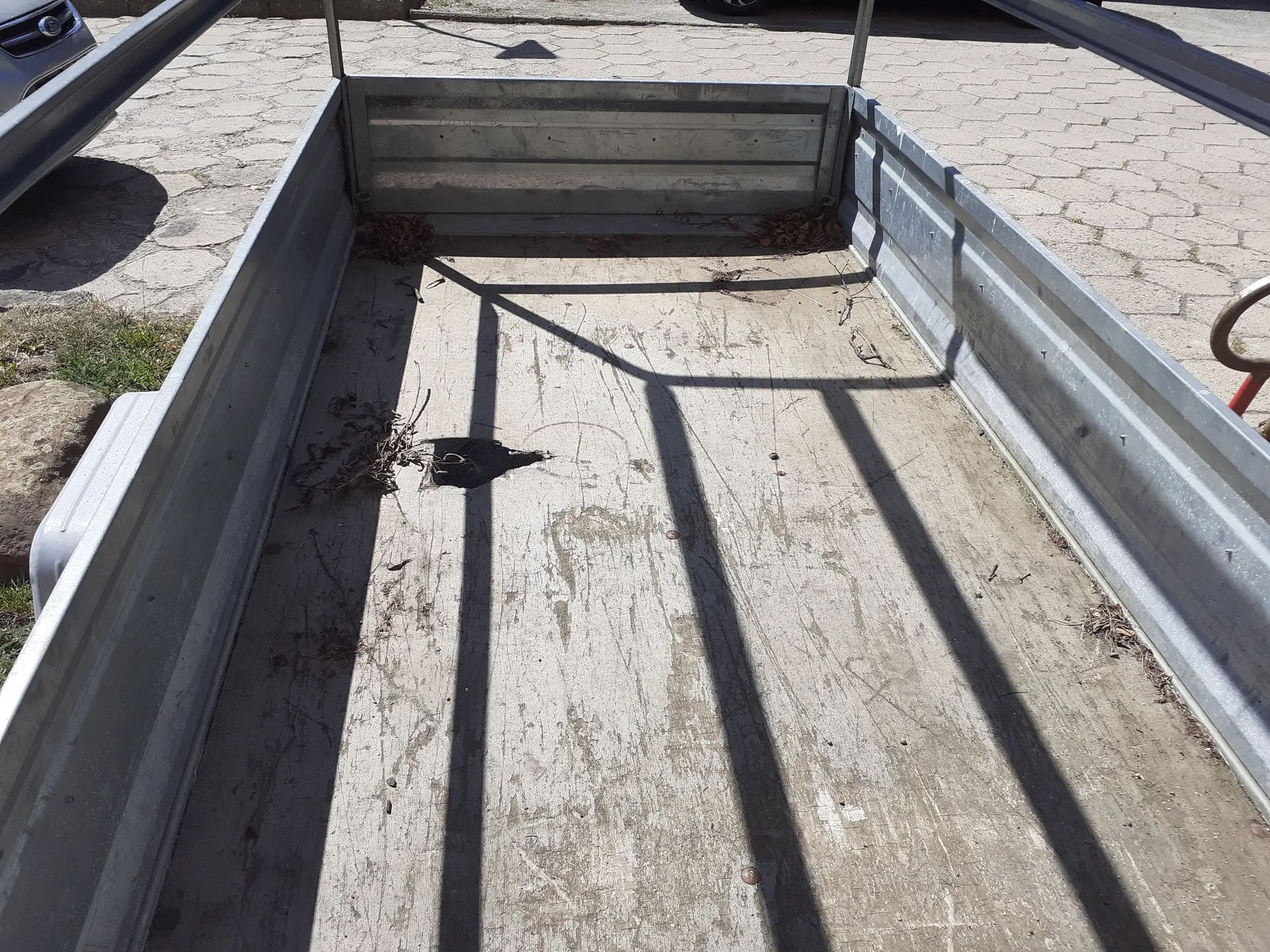 Widok z góry do wnętrza metalowej przyczepy transportowej. Przyczepa ma ocynkowane ściany i drewnianą podłogę, na której znajdują się resztki liści i gałązek. Na dnie widoczne są wyraźne, równoległe cienie rzucane przez konstrukcję nad przyczepą. W tle widać fragmenty chodnika z kostki brukowej oraz fragment srebrnego samochodu po lewej stronie.