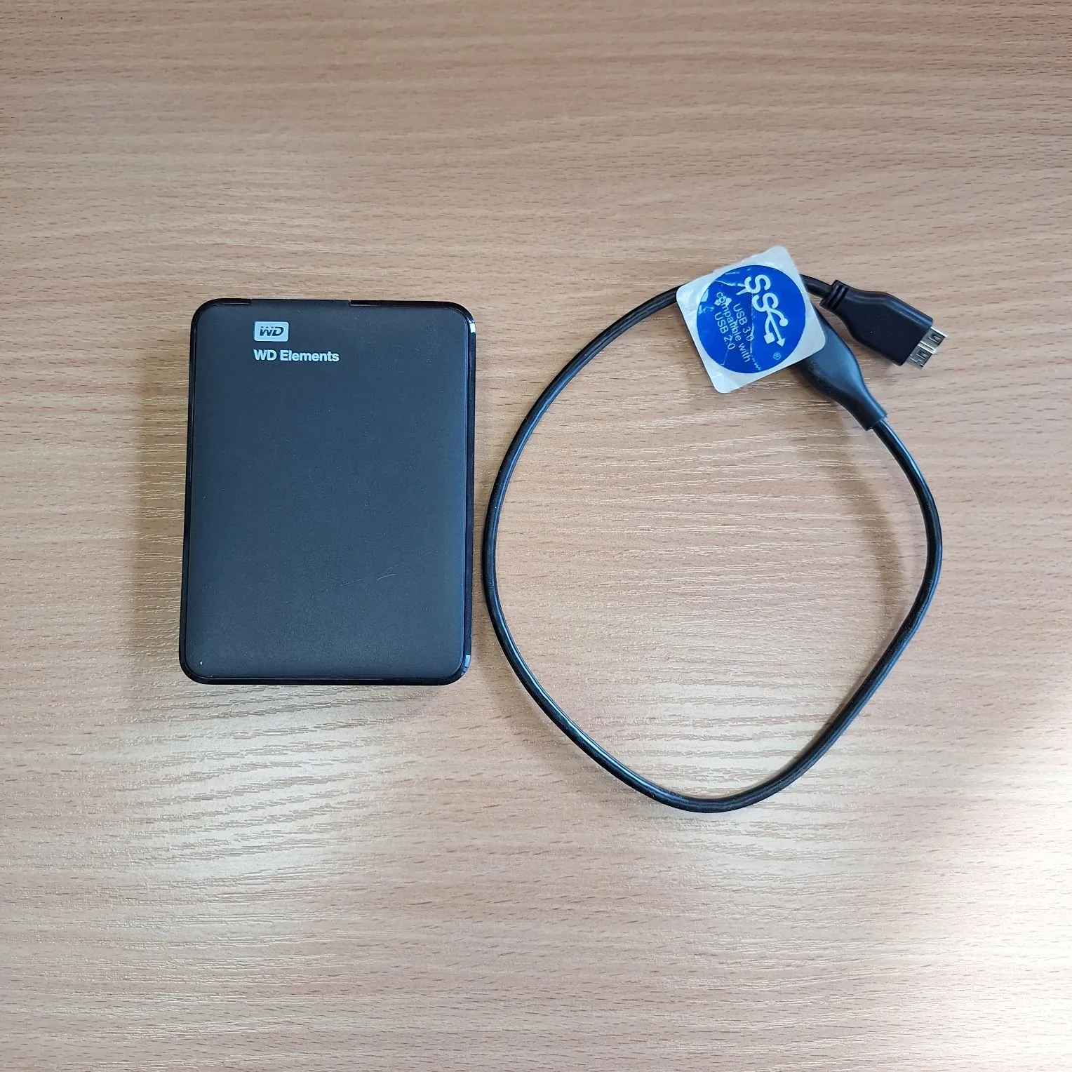 Czarny zewnętrzny dysk twardy marki WD z napisem 'WD Elements' oraz dołączony do niego czarny kabel USB z niebieską etykietą, leżące na jasnym, drewnianym blacie.