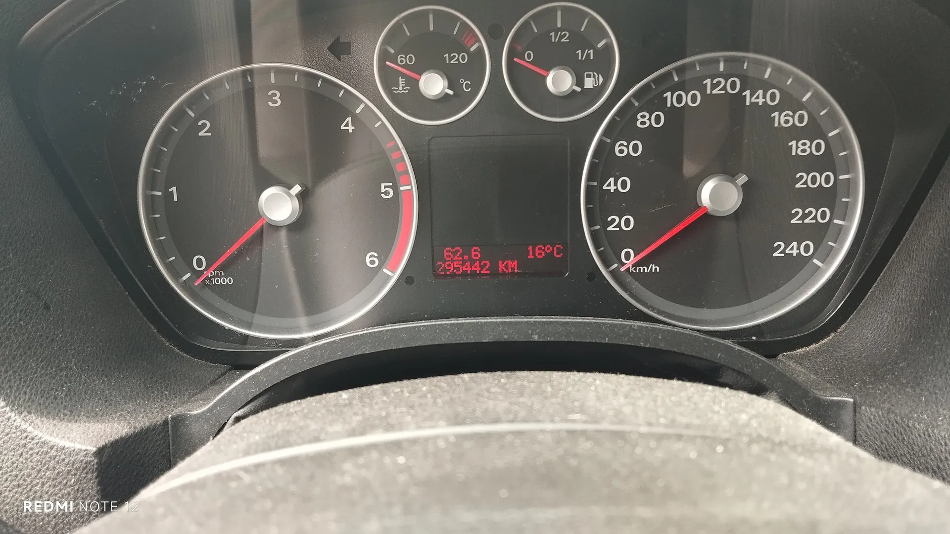 Tablica przyrządów samochodowych z tachometrem (0-6 x1000 obr./min), szybkościomierzem (0-240 km/h), wskaźnikiem temperatury chłodniczki (0-120°C) i paliwa (0-1/2). W centralnym wyświetlaczu cyfrowym widoczne wartości: 62.6, 16°C oraz 295442 KM. Na dole obrazu widoczna część kierownicy. W lewym dolnym rogu znak wodny 'REDMI NOTE 12'.