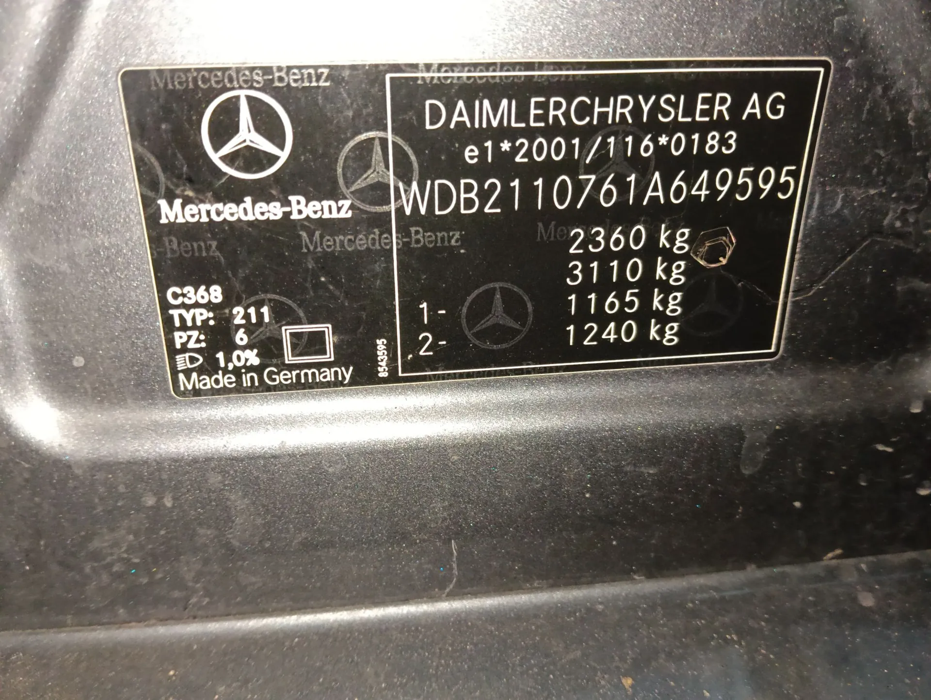 Tabliczka znamionowa samochodu marki Mercedes-Benz. Po lewej stronie znajduje się logo gwiazdy oraz napis 'Mercedes-Benz'. Po prawej stronie widnieje tekst: 'DAIMLERCHRYSLER AG', numer homologacji 'e1*2001/116*0183' oraz numer VIN 'WDB2110761A649595'. Poniżej znajdują się dane techniczne: 'C368', 'TYP: 211', 'PZ: 6', '1,0%', 'Made in Germany' oraz wartości masy: '2360 kg', '3110 kg', '1165 kg', '1240 kg'.