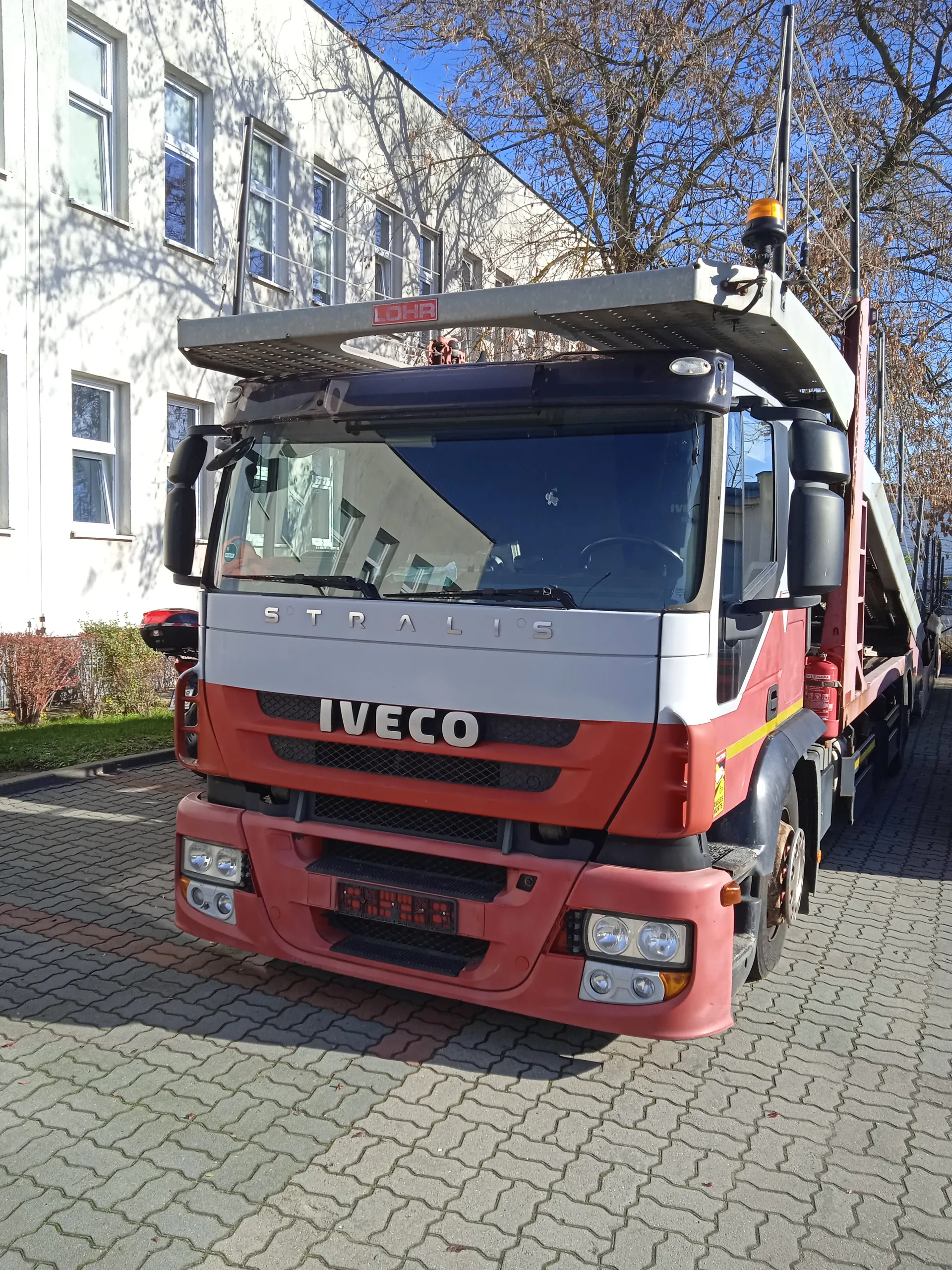 Czerwono-biały ciężarówka Iveco Stralis z napisem 'LOHR' na dachu, zaparkowana na brukowanej powierzchni. W tle biały budynek z oknami, drzewa bez liści i jasne niebo. Na przodzie pojazdu widoczne napisy 'STRALIS' i 'IVECO'.