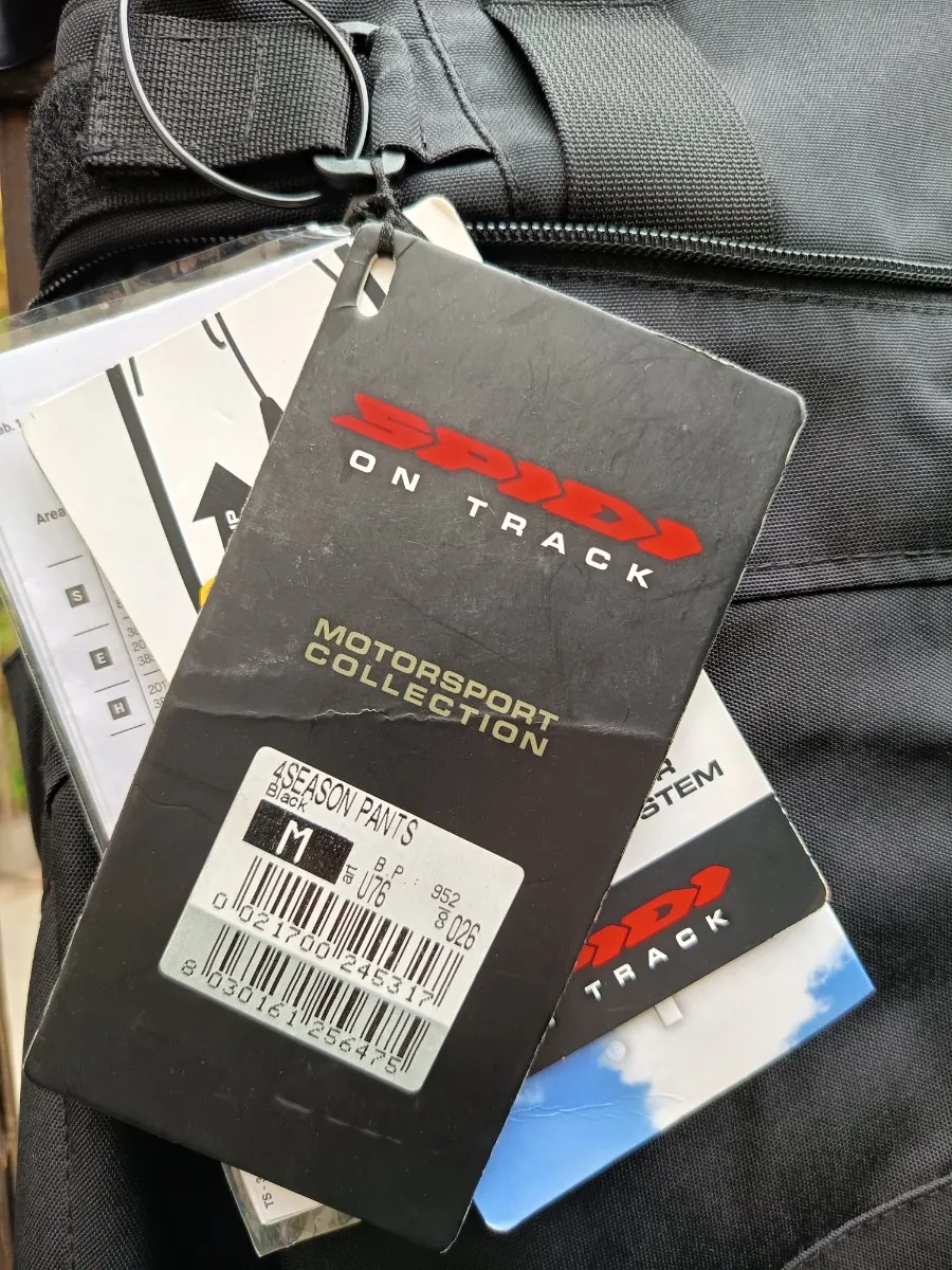 Zbliżenie na czarną metkę produktu marki Spidi, przymocowaną do czarnego materiału z nylonu. Na metce widnieje czerwone logo 'SPIDI', biały napis 'ON TRACK' oraz 'MOTORSPORT COLLECTION'. W dolnej części metki znajduje się opis produktu: '4SEASON PANTS', kolor 'Black', rozmiar 'M' oraz kody kreskowe z numerami seryjnymi. Pod główną metką widoczne są inne etykiety i instrukcje.