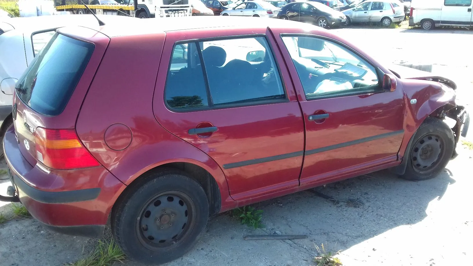 Czerwony samochód osobowy marki Volkswagen Golf w nadwoziu kombi, zaparkowany na placu. Pojazd jest uszkodzony w przedniej części po prawej stronie, gdzie widać wgniecioną karoserię i brak przedniego zderzaka. Samochód ma czarne stalowe felgi i czarne listwy boczne.