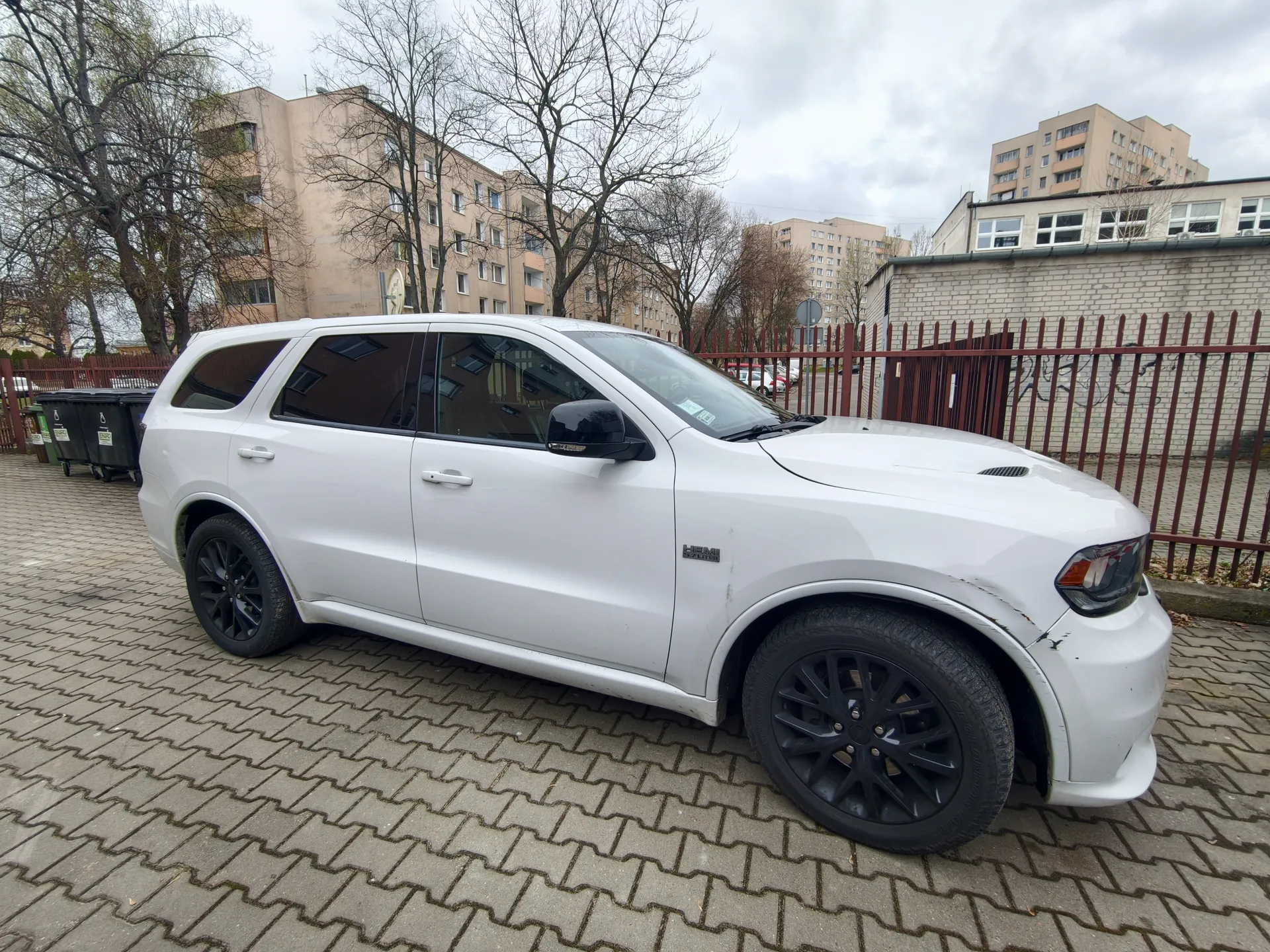 Biały SUV Dodge Durango SRT z czarnymi felgami zaparkowany na brukowanej powierzchni. Widoczne uszkodzenie przedniego zderzaka. W tle bloki mieszkalne, drzewa bez liści, czerwone ogrodzenie i pojemniki na śmieci. Niebo zachmurzone.