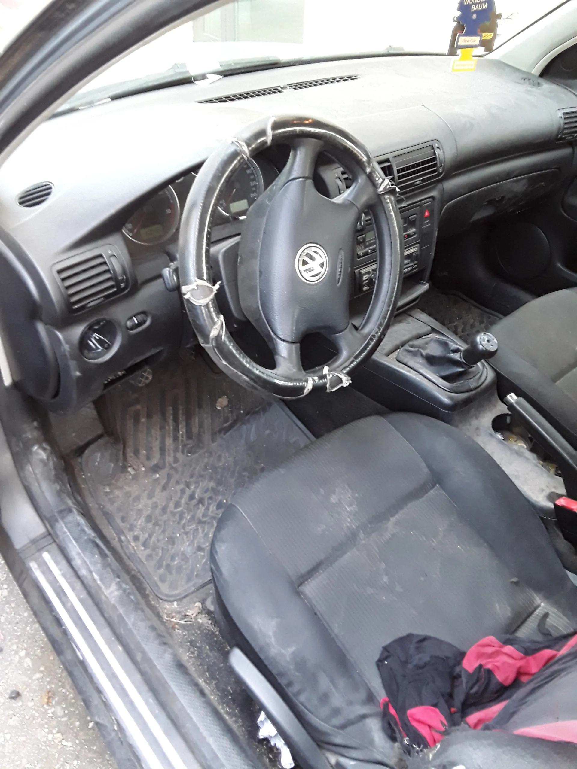 Wnętrze samochodu z widocznym kierownicą marki Volkswagen, czarnym siedzeniem pasażera z czerwono-czarną kurtką, przyciskami sterowania na desce rozdzielczej, dźwignią zmiany biegów oraz brudnymi dywanikami podłogowymi. Na lewej stronie widoczny jest niebieski znaczek z częściowo czytelnym tekstem.