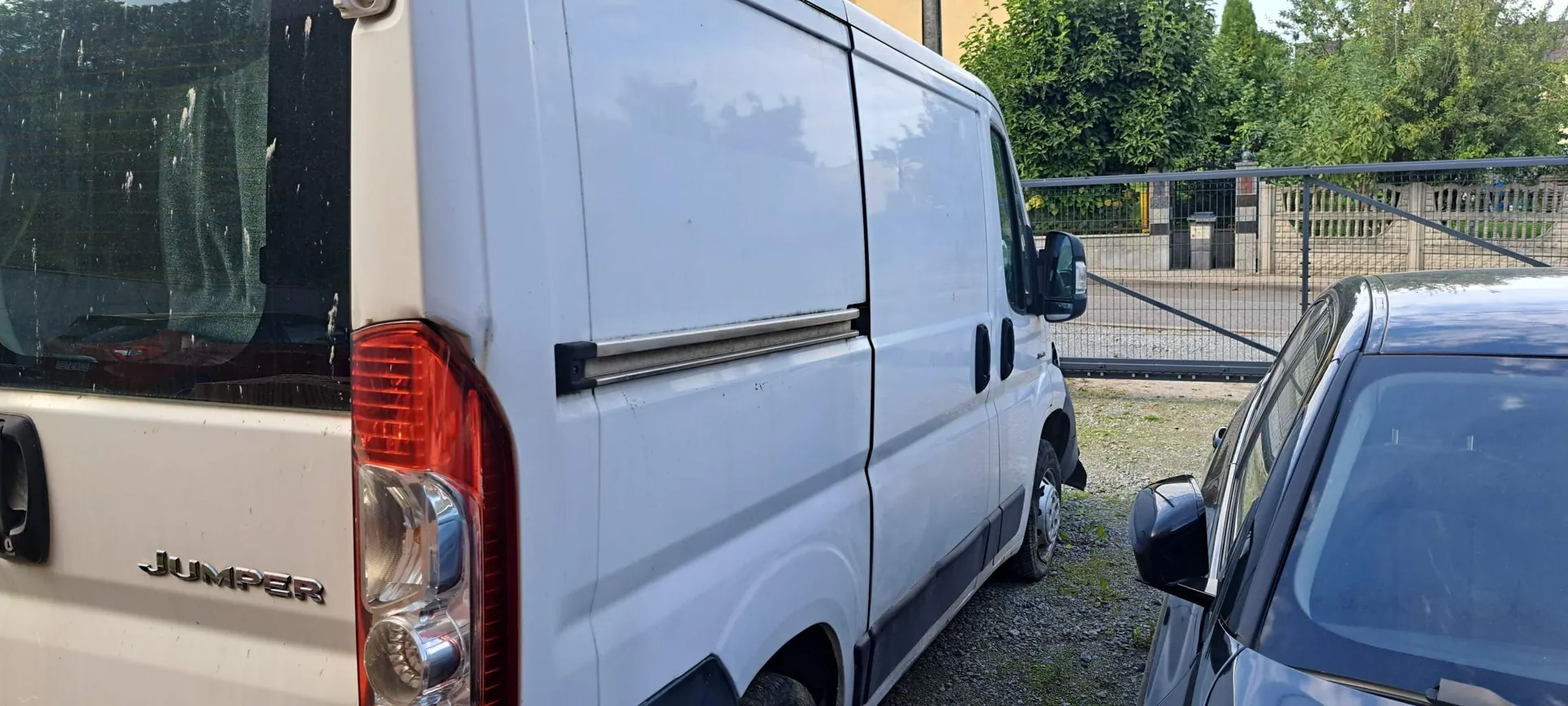Zbliżenie na tył i prawy bok białego vana marki Citroën Jumper. Na klapie bagażnika widoczny jest napis 'JUMPER'. Obok vana zaparkowany jest ciemny samochód osobowy. W tle widać metalowy płot oraz zielone drzewa.
