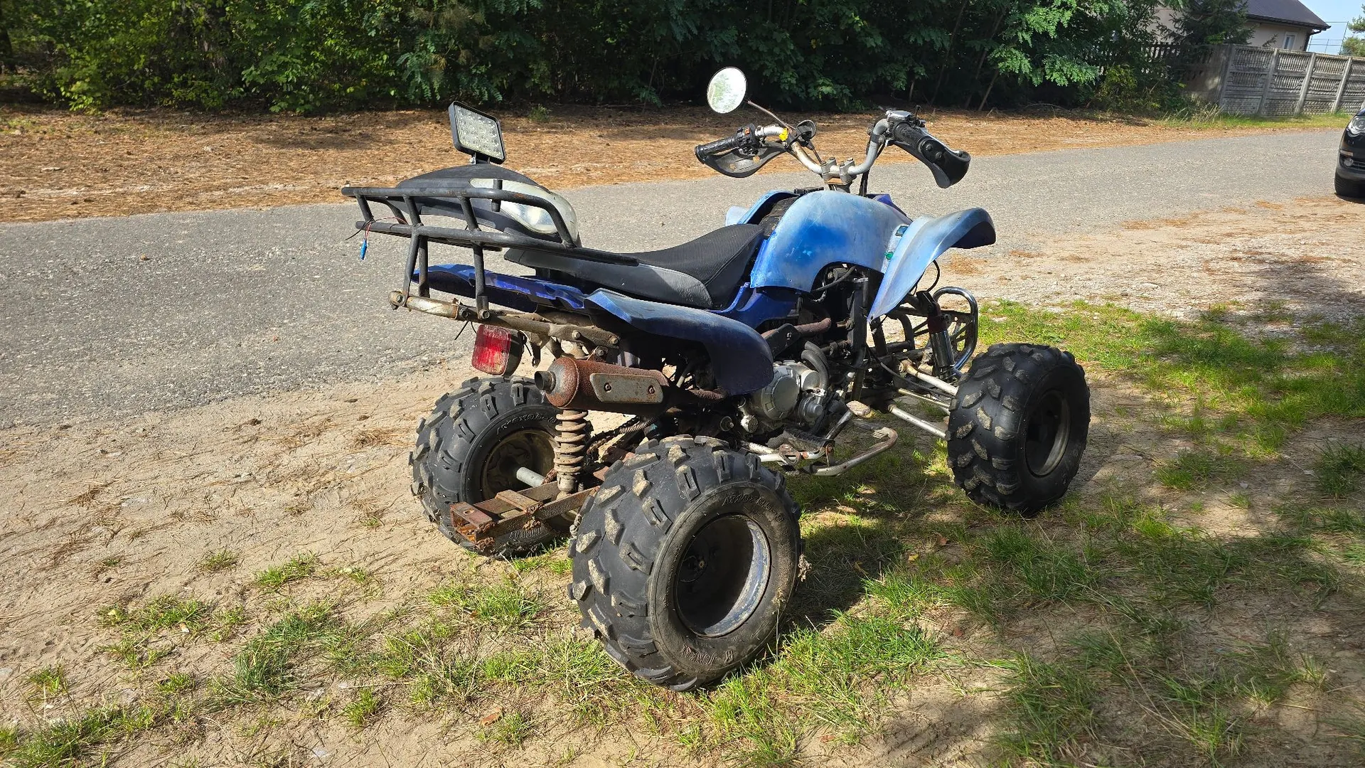 Niebieski quad zaparkowany na piaszczysto-trawiastej drodze. Pojazd posiada czarne siedzenie, metalowy bagażnik z tyłu, lusterka boczne oraz duże, terenowe opony z wyraźnym bieżnikiem. W tle widoczna jest zielona roślinność i fragment ogrodzenia.