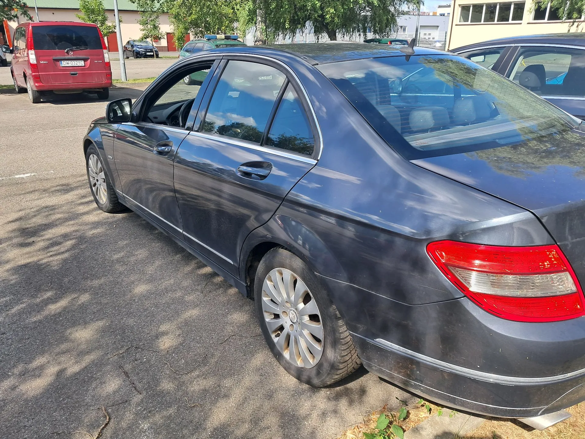 Srebrno-niebieski samochód osobowy marki Mercedes-Benz zaparkowany na asfaltowym placu. Auto jest pokazane z profilu od strony pasażera, z widocznym tyłem i prawym bokiem. W tle widoczny jest czerwony samochód dostawczy z tablicą rejestracyjną DW 032YQ oraz inne zaparkowane pojazdy i budynki przemysłowe.