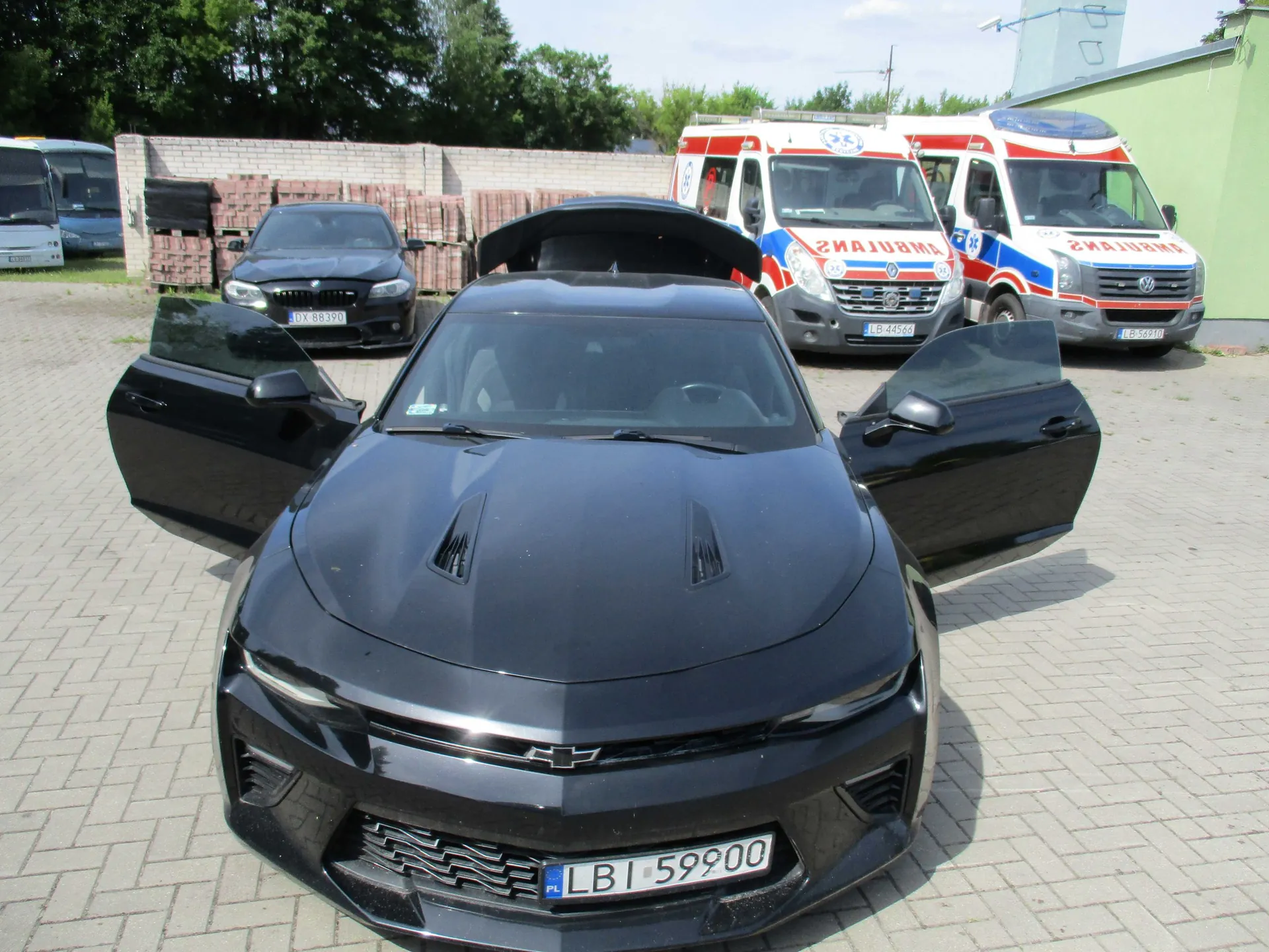 Czarny samochód sportowy marki Chevrolet z otwartymi drzwiami i otwartym schowkiem nad przednią szybą, zaparkowany na kostce brukowej. Samochód ma polską tablicę rejestracyjną LBI 59900. W tle widoczne są dwa białe ambulanse z napisami 'RATOWNICTWO' oraz ciemny samochód osobowy z tablicą DX 8839D. W oddali znajduje się mur z cegieł i zielone drzewa.