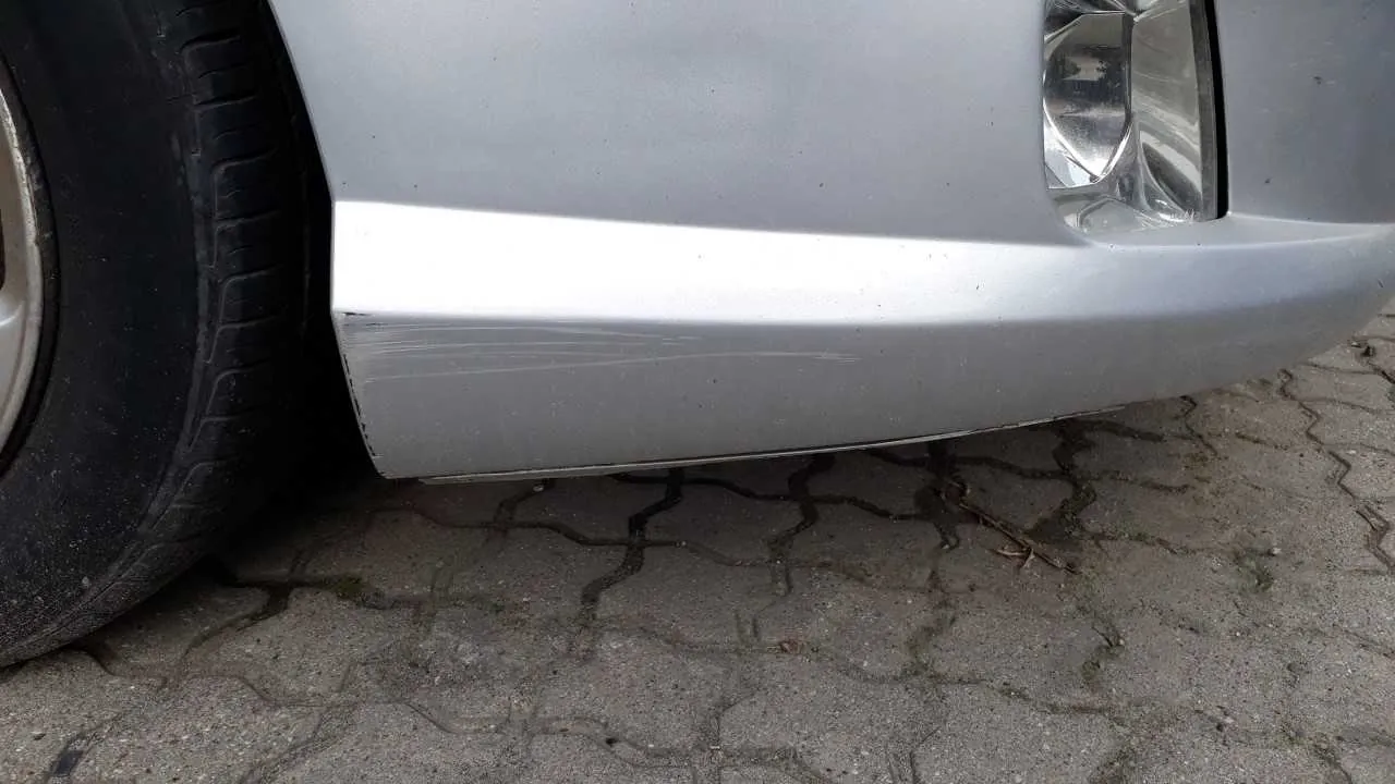 Przednia część srebrnego samochodu z widocznymi zadrapaniami na zderzaku. Po lewej stronie widoczne jest przednie koło z oponą, po prawej reflektor. Samochód stoi na brukowanej powierzchni z kamiennymi płytami ułożonymi w geometryczny wzór. Brak czytelnego tekstu.