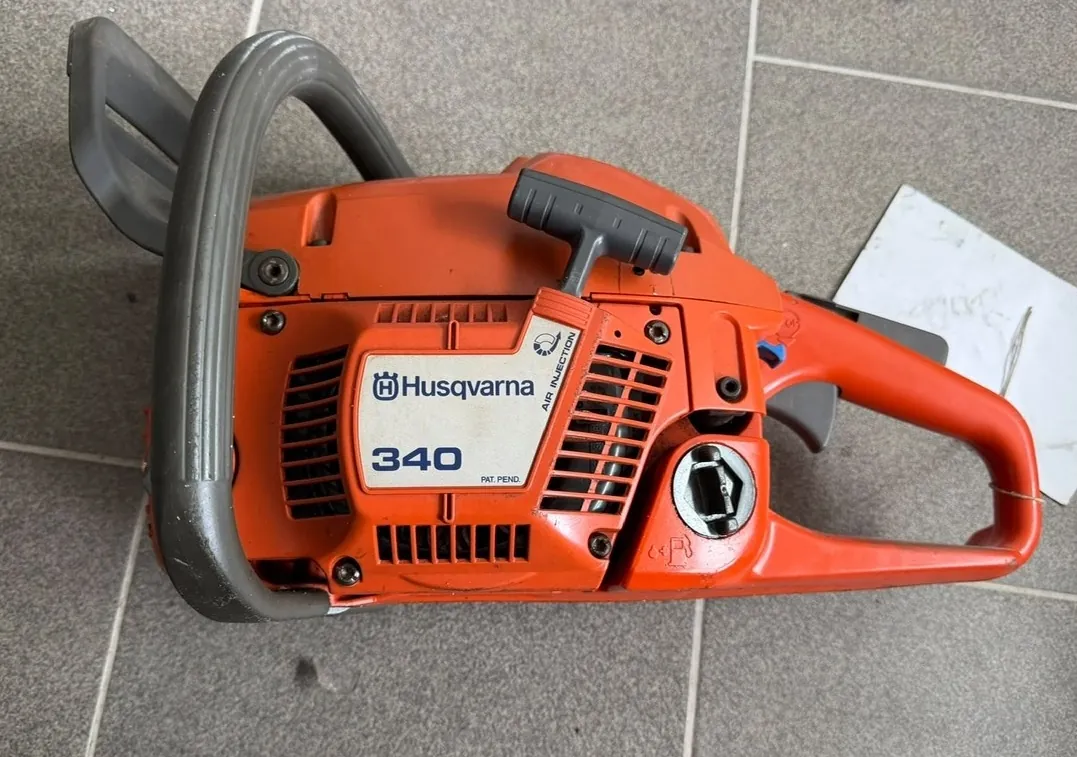 Pomarańczowa piła spalinowa marki Husqvarna, model 340, leżąca na szarych płytkach ceramicznych. Na obudowie widnieje napis 'Husqvarna 340' oraz mniejszy napis 'AIR INJECTION' i 'PAT. PEND.'. Urządzenie posiada szary uchwyt oraz czarną rączkę rozrusznika.