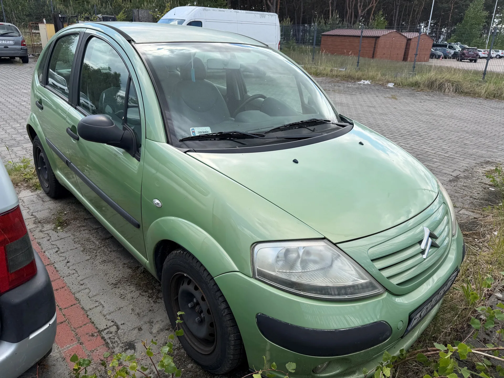 Zielony samochód osobowy marki Citroën zaparkowany na brukowanym placu. Widoczne są czarne elementy nadwozia, reflektory, logo marki na masce oraz tablica rejestracyjna. W tle biały furgon, czerwone budki, ogrodzenie, drzewa i inne zaparkowane pojazdy. Na przedniej szybie naklejka. Samochód znajduje się na obszarze z trawą i roślinnością przy krawędzi parkingu.