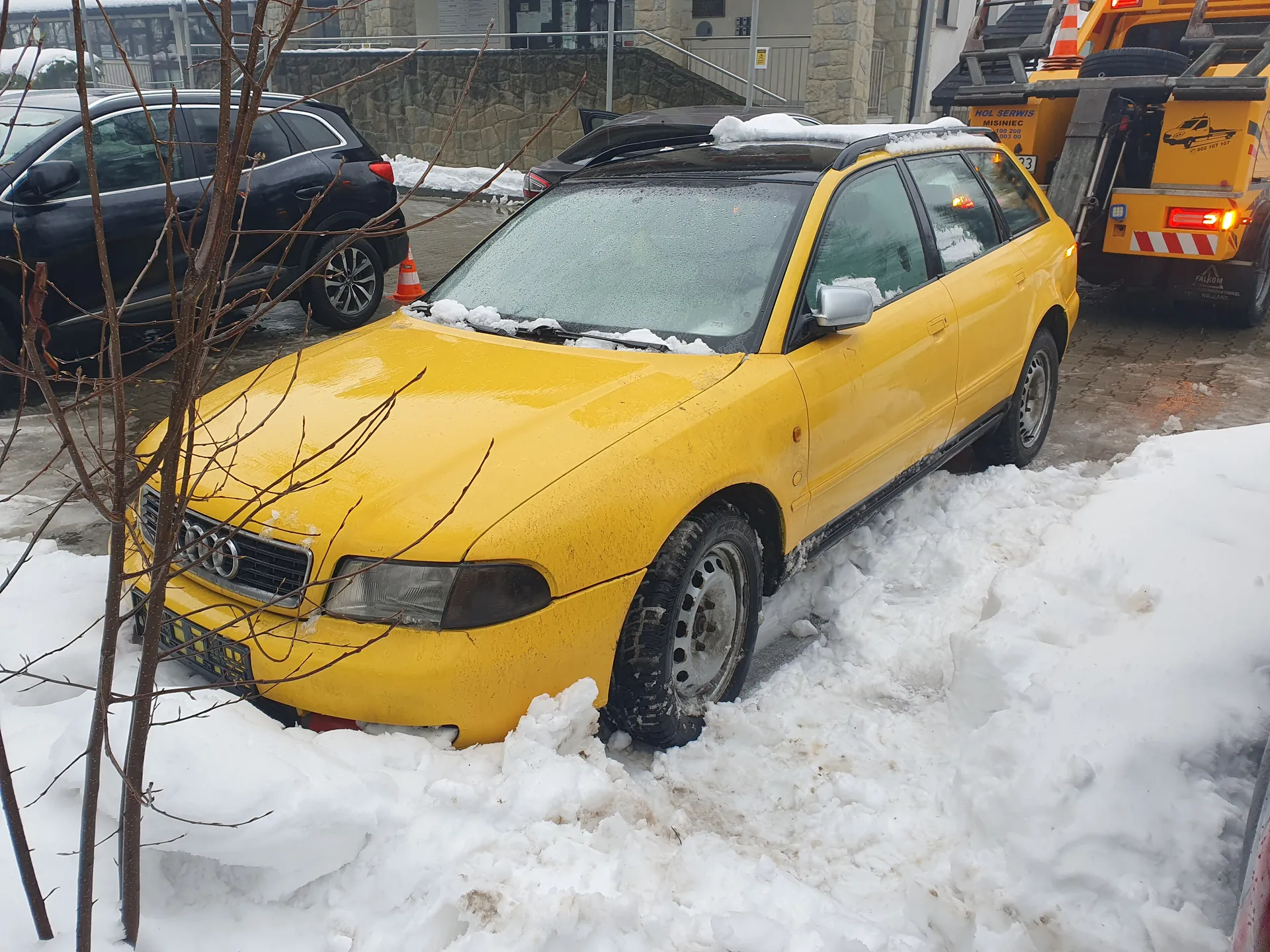 żółty samochód Audi kombi pokryty śniegiem, zaparkowany na śnieżnym placu parkingowym. W tle czarny SUV, żółty wóz holowniczy z napisem "HOL SERWIS MISINIEC" oraz kamienica z kamienną ścianą i schodami. Na pierwszym planie bezlistne drzewko. Śnieg na ziemi i na dachach pojazdów.