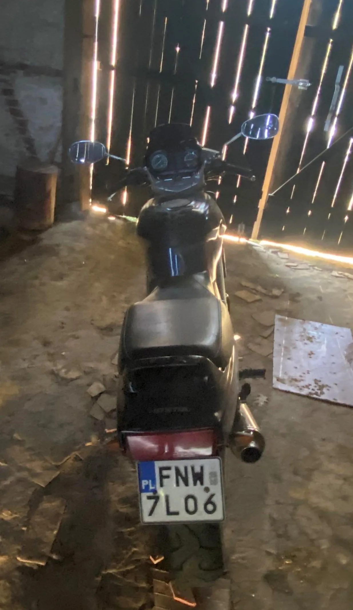 Czarny motocykl widziany od tyłu, stojący na brudnej podłodze z rozrzuconymi kawałkami materiału. Widoczna tablica rejestracyjna z napisem PL FNW 7L06, czerwone światło tylnego reflektora oraz dwa lusterka boczne. W tle pionowe drewniane belki i betonowa ściana po lewej stronie. Oświetlenie tworzy poziome pasy światła w tle.