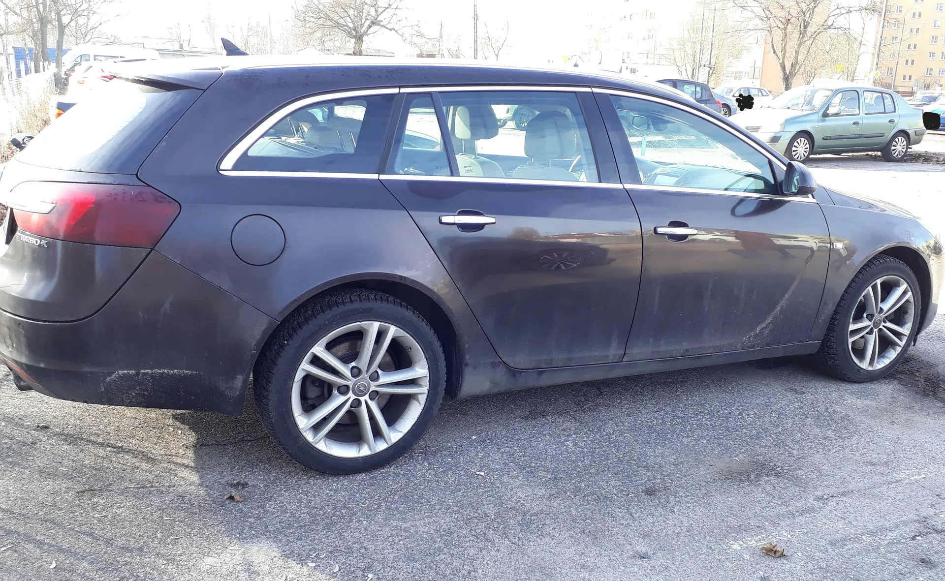 Ciemnoszary samochód typu kombi zaparkowany na asfaltowej powierzchni. Pojazd jest zabrudzony, widoczne są srebrne felgi i napis TURBO na tylnej części. W tle inne samochody, drzewa bez liści oraz budynki mieszkalne. Słońce oświetla scenę, rzucając cienie na ziemię.