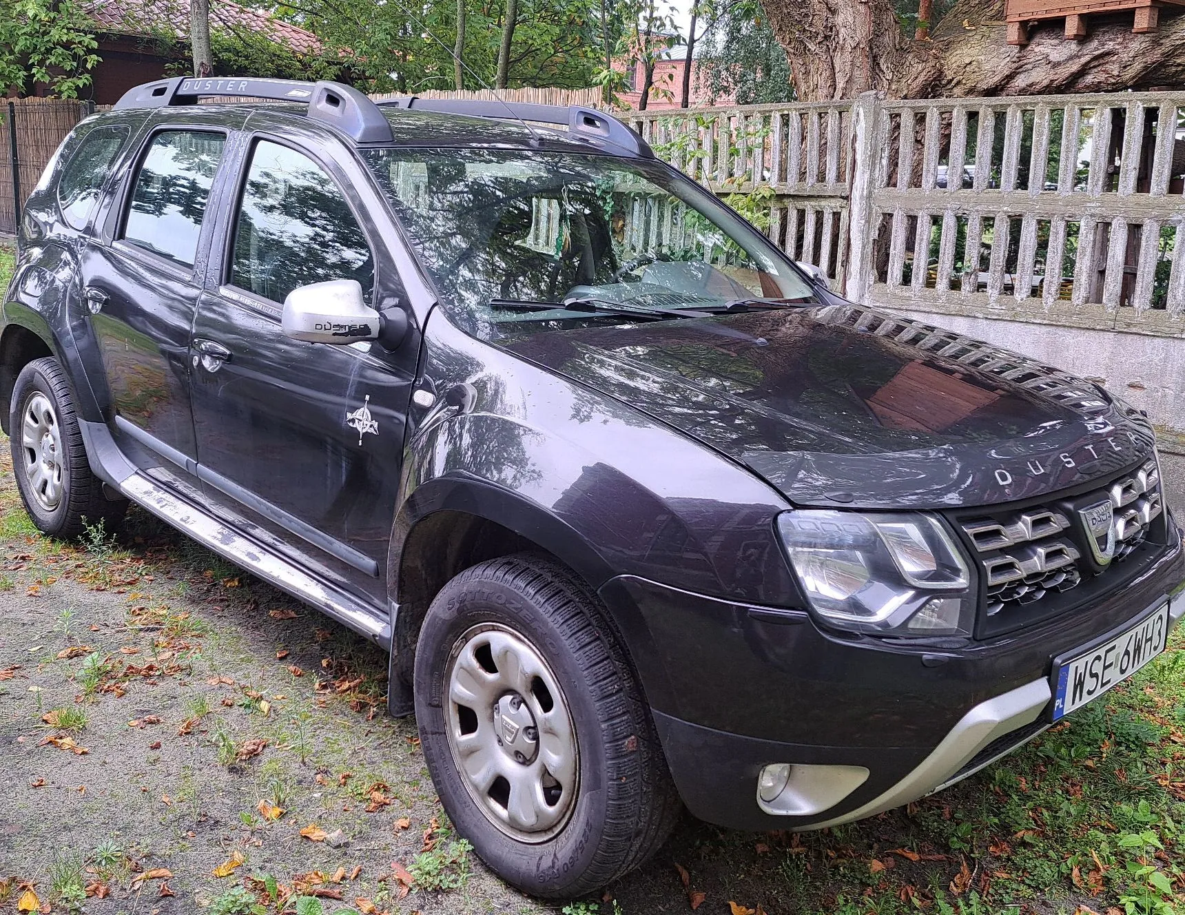 Ciemnoszary samochód marki Dacia Duster zaparkowany na trawiastym podłożu obok drewnianego ogrodzenia. Pojazd posiada srebrne relingi dachowe, stopnie boczne oraz tablicę rejestracyjną o numerze WSE 6WH3.