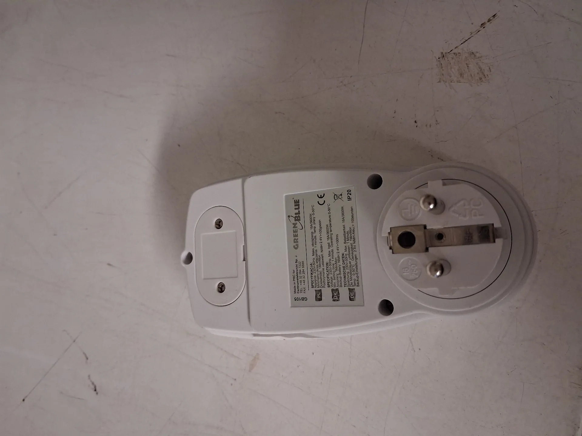 Zdjęcie białego adaptera elektrycznego leżącego na jasnej powierzchni. Na obudowie urządzenia znajduje się logo 'GREEN BLUE' oraz szczegółowe dane techniczne i certyfikaty, w tym oznaczenia CE i IP20. Po prawej stronie widoczne jest gniazdo z dwoma pinami oraz otwór na wtyczkę.
