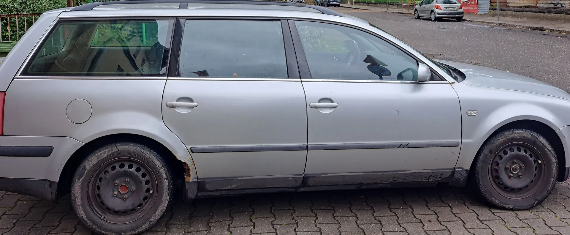 Srebrny samochód typu kombi zaparkowany na kostce brukowej. Widoczny jest profil prawy pojazdu z czarnymi stalowymi felgami. Na dolnej krawędzi nadwozia, za przednim kołem, widoczne są ślady korozji i rdzy.