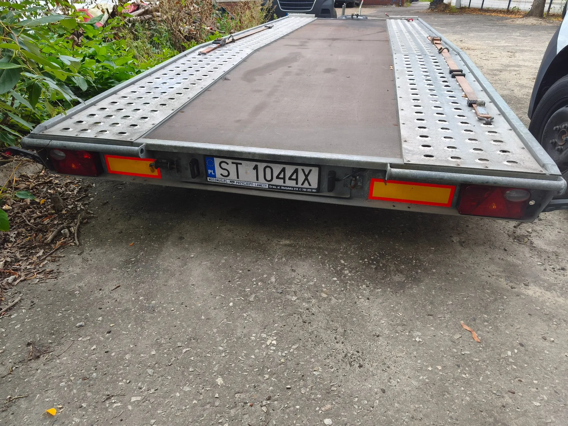 Widok z tyłu na metalową lawetę samochodową z platformą transportową. Platforma składa się z centralnej, ciemnej części antypoślizgowej oraz bocznych pasów z perforacją. Na tylnej belce znajduje się polska tablica rejestracyjna o numerze ST 1044X, dwa czerwone odblaski oraz światła tylne. Laweta stoi na szutrowym podłożu, w tle widoczna jest roślinność.