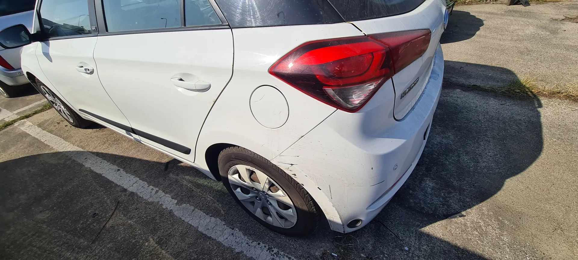 Biały samochód osobowy typu hatchback zaparkowany na asfaltowym parkingu. Widok z tyłu i boku. Czerwone światło tylnego reflektora, srebrne felgi, pokrywa zbiornika paliwa po lewej stronie. Na tylnym zderzaku widoczne zadrapania. Biały znak parkingu na asfalcie. W tle częściowo widoczny inny srebrny samochód. Cień pojazdu na powierzchni parkingu.