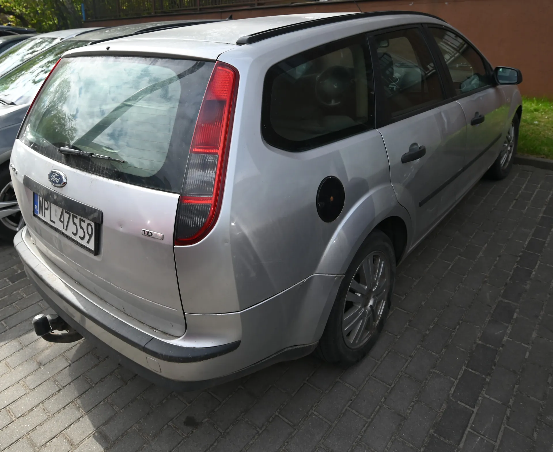 Srebrny samochód marki Ford, widok z tyłu i boku. Widoczna tablica rejestracyjna WPL 47559, logo Ford oraz oznaczenie TD na tylnej części pojazdu. Czerwone światła tylne, czarne okna i czarna obudowa lusterka bocznego. Samochód zaparkowany na brukowanej powierzchni, w tle częściowo widoczne inne pojazdy, brązowa ściana budynku i zielona trawa.