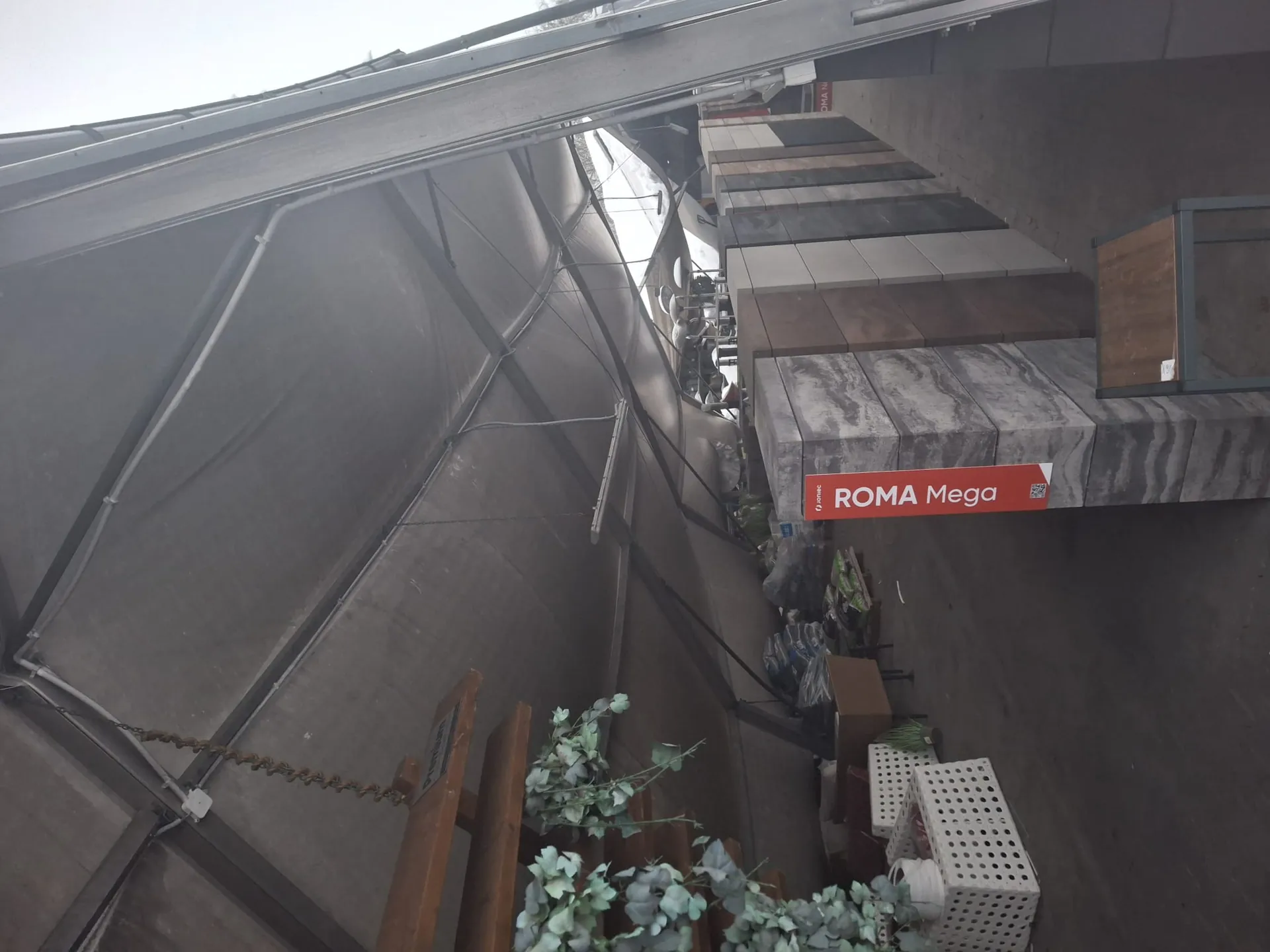 Zdjęcie przedstawia przestrzeń pod namiotem lub dachem z szarym materiałem. Po lewej stronie widoczne są drewniane półki z sztucznymi roślinami oraz białe kratowane pudełka. W centrum ustawione są wysokie bloki z różnymi teksturami (marmurowa, drewniana, betonowa). Na czerwonym pionowym znaku znajduje się biały tekst: "ROMA Mega" oraz mniejszy napis "Fornir" u góry. W tle widoczne są worki z materiałami i meble. Podłoga jest betonowa, a po prawej stronie znajduje się chodnik.