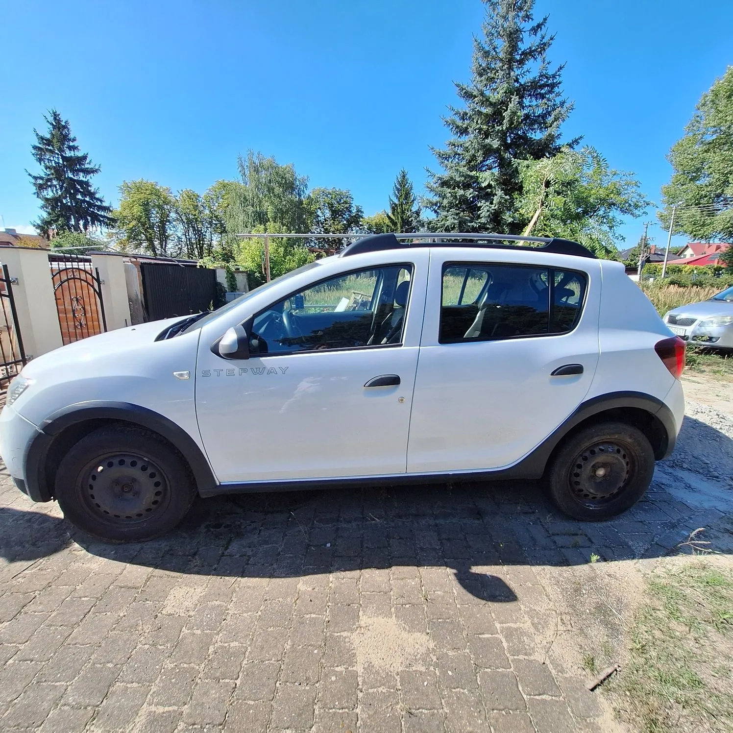 Biały samochód marki Dacia Stepway zaparkowany bokiem na kostce brukowej. Na przednich drzwiach widnieje napis 'STEPWAY'. Pojazd posiada czarne plastikowe osłony nadkoli i progi. W tle widać zielone drzewa, niebieskie niebo oraz fragment ogrodzenia i budynków.