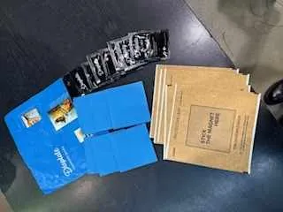 Na ciemnym stole leżą trzy grupy przedmiotów: niebieski folder z widocznym napisem 'Szybka' i małym zdjęciem, stos brązowych kopert z napisem 'Small Package' oraz czarno-biały pasek z nieczytelnym tekstem. W tle widoczny jest fragment podłogi.